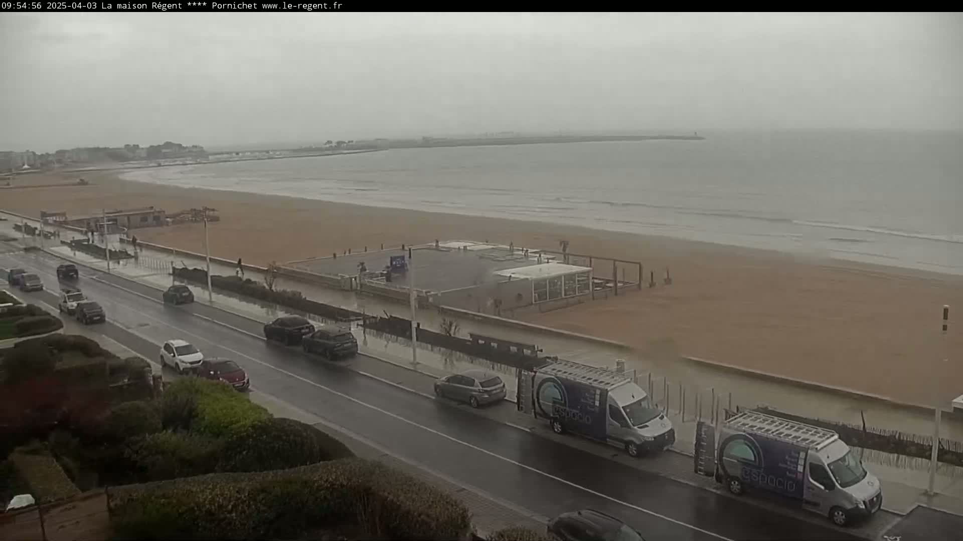 A rain-swept beach scene shows a road with several cars parked along the side, next to a sandy beach with gentle waves and a building on the shore.