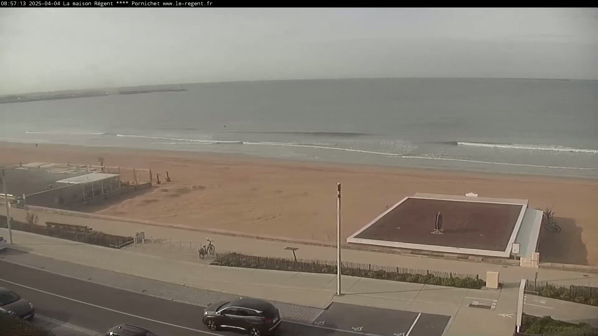 An overcast day at a beach shows a sandy shore with small waves, a beach volleyball court, some cars parked along a road, and a building near the water.