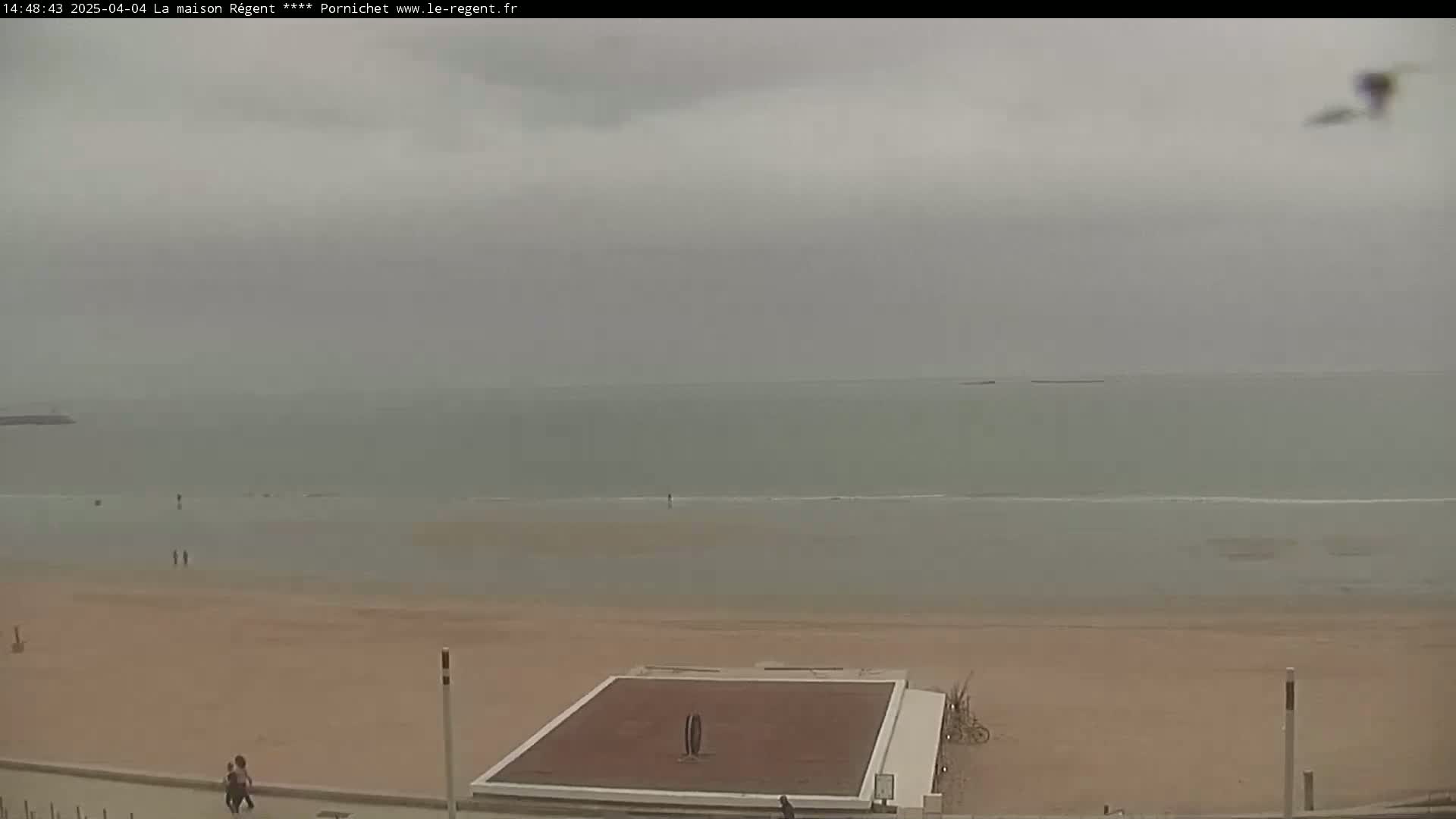 A mostly cloudy day at a beach shows a calm sea, a wide sandy shore, and a few people walking near a paved area with a large flat structure.