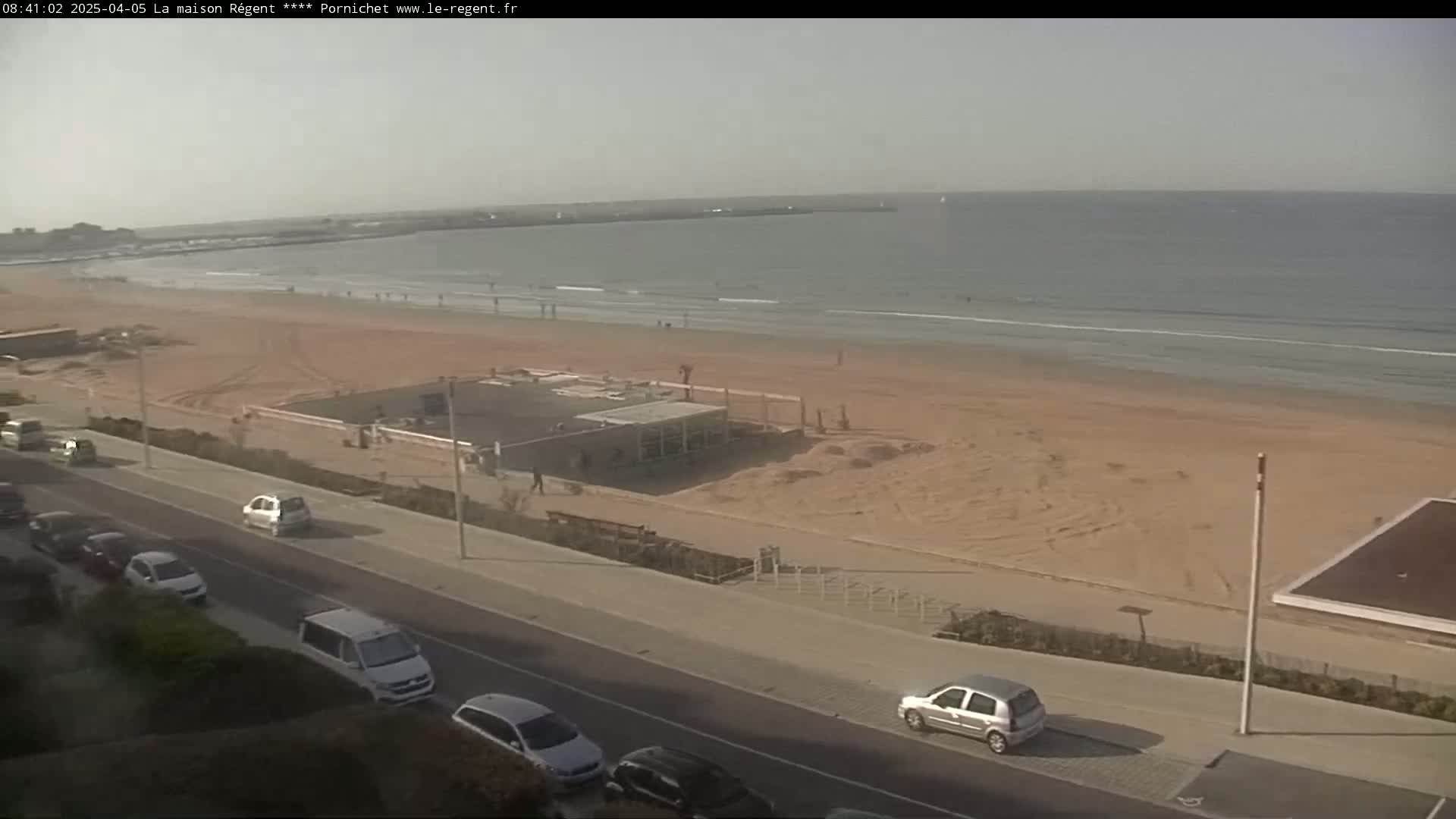 A sandy beach with a calm ocean and several cars parked along a road next to it under a hazy sky.
