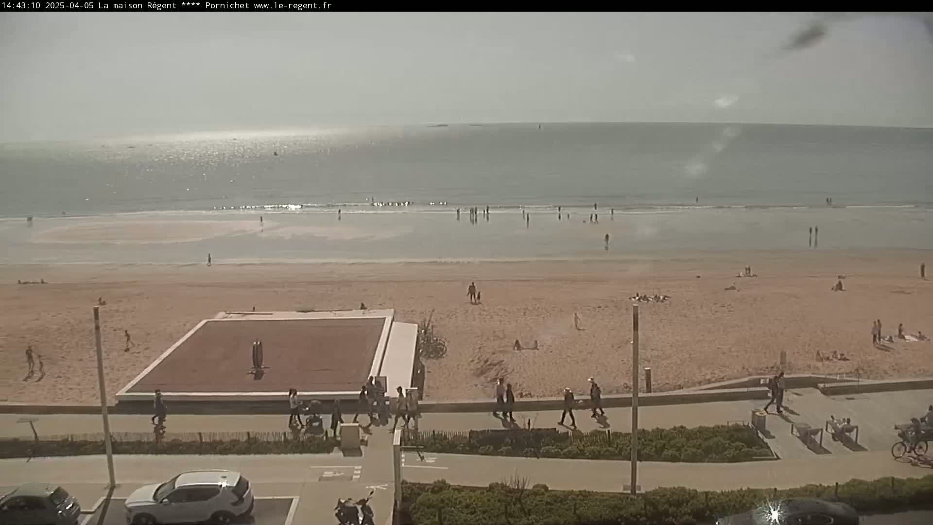 A beach scene on a sunny day shows a calm ocean with several people in the water and on the sandy shore, near a paved walkway and parking lot.