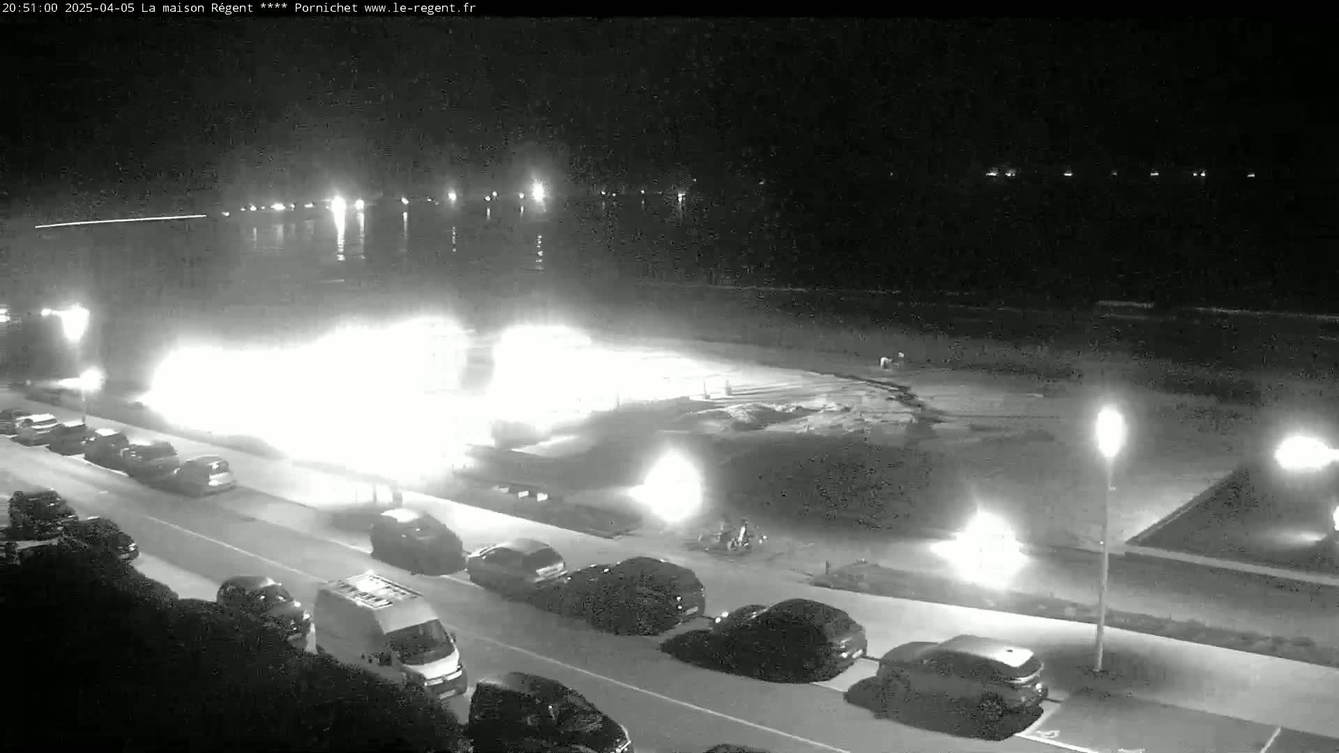 A nighttime, black-and-white view of a street with parked cars alongside a beach and body of water, illuminated by streetlights.