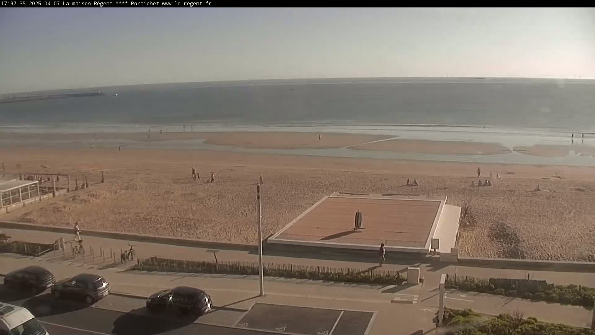 A sandy beach with a few people on it stretches to a calm ocean under a clear sky.
