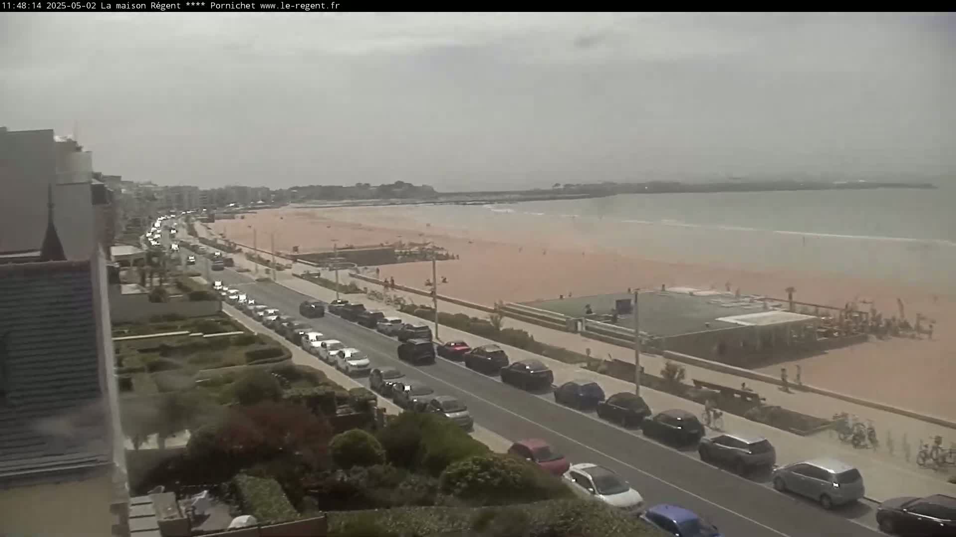 A long road lined with parked cars runs alongside a sandy beach and calm ocean under an overcast sky.