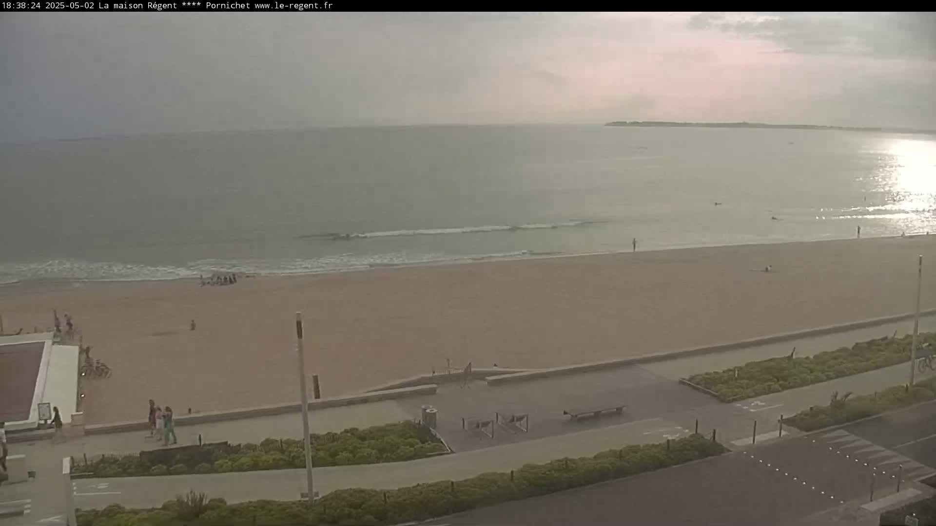 A mostly sunny day shows a wide sandy beach with a few people and small waves lapping the shore, alongside a paved walkway and green landscaping.