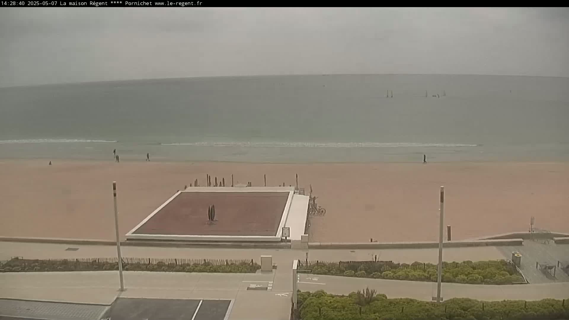 A mostly overcast day reveals a sandy beach with a few people and a reddish-brown paved court near the water's edge, a calm ocean in the background.