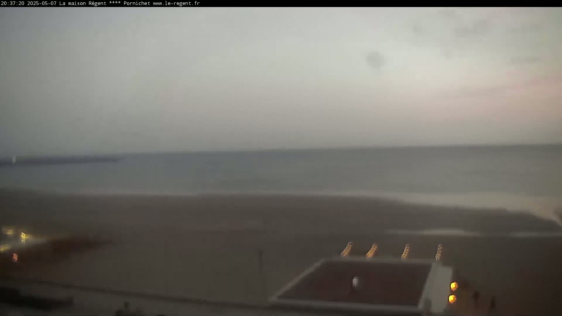 A hazy, overcast view shows a beach with a calm ocean in the background and some lights near a structure on the sand.