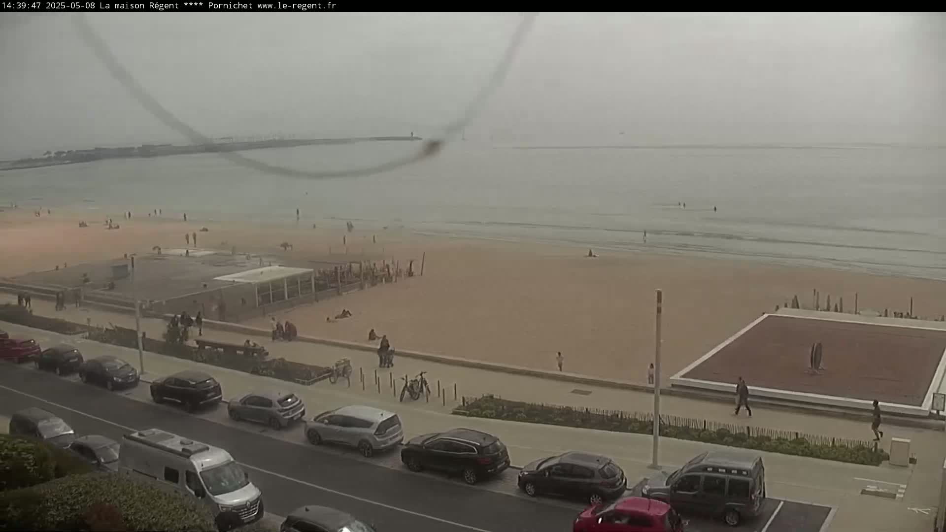 A hazy, overcast day shows a sandy beach with scattered people, a boardwalk lined with parked cars, and a calm ocean in the background.
