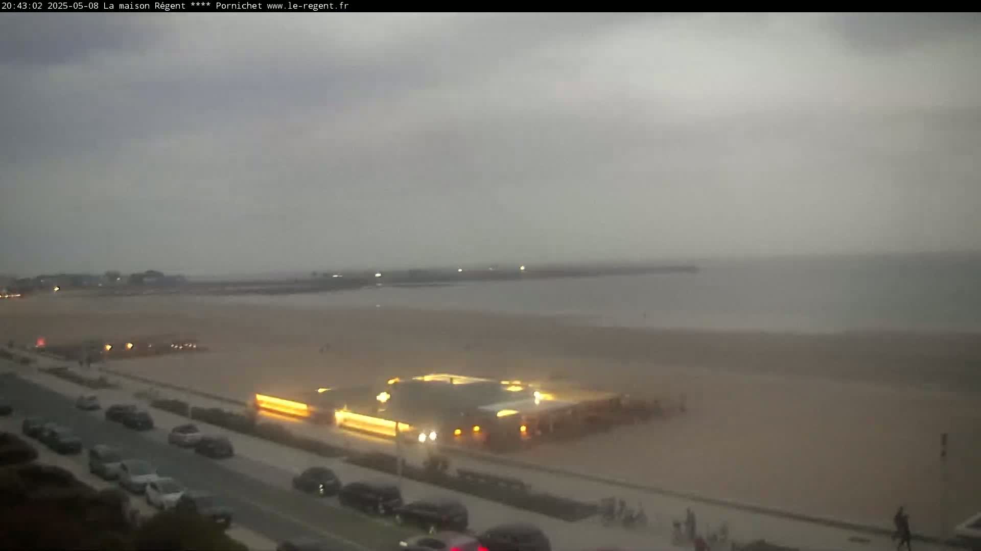 An overcast evening scene shows a beach with a lit-up beachfront building, cars parked along a road, and a few people walking near the water's edge.