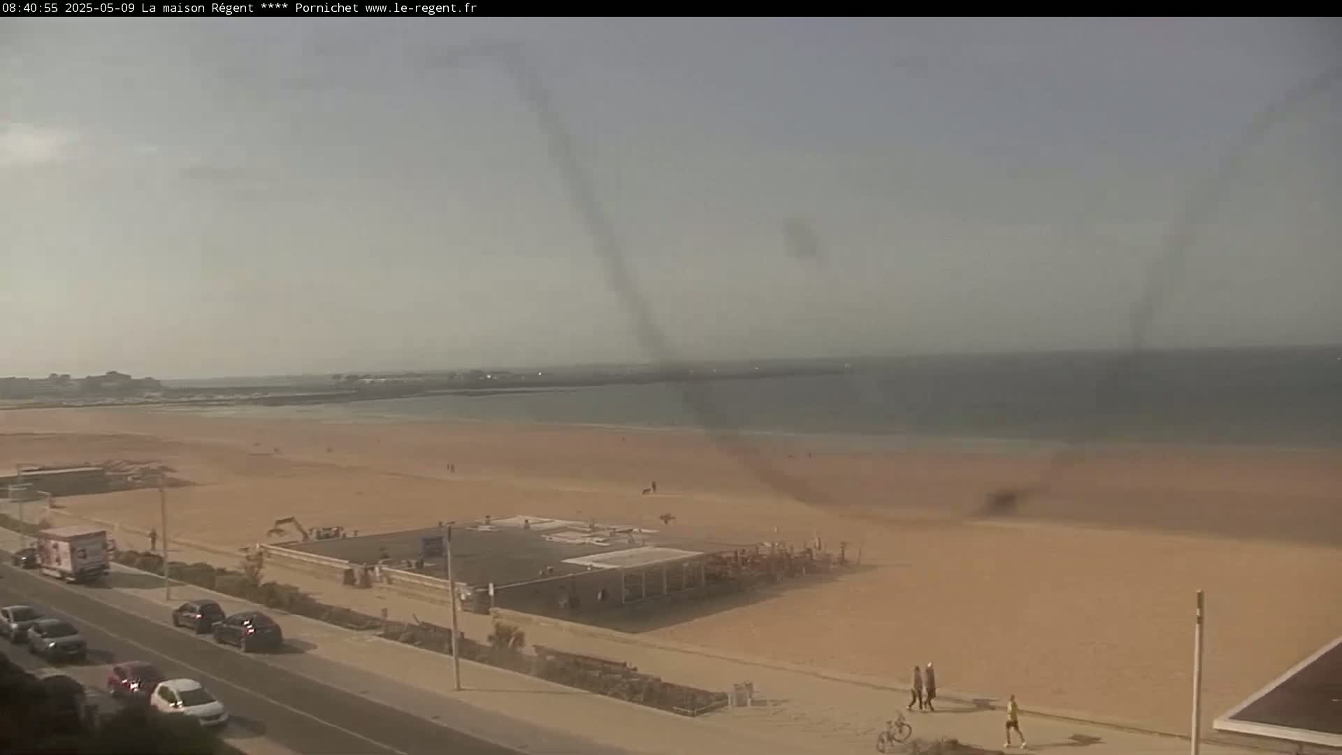 A hazy, sunny day overlooks a wide sandy beach with a few people and vehicles parked along a nearby road.