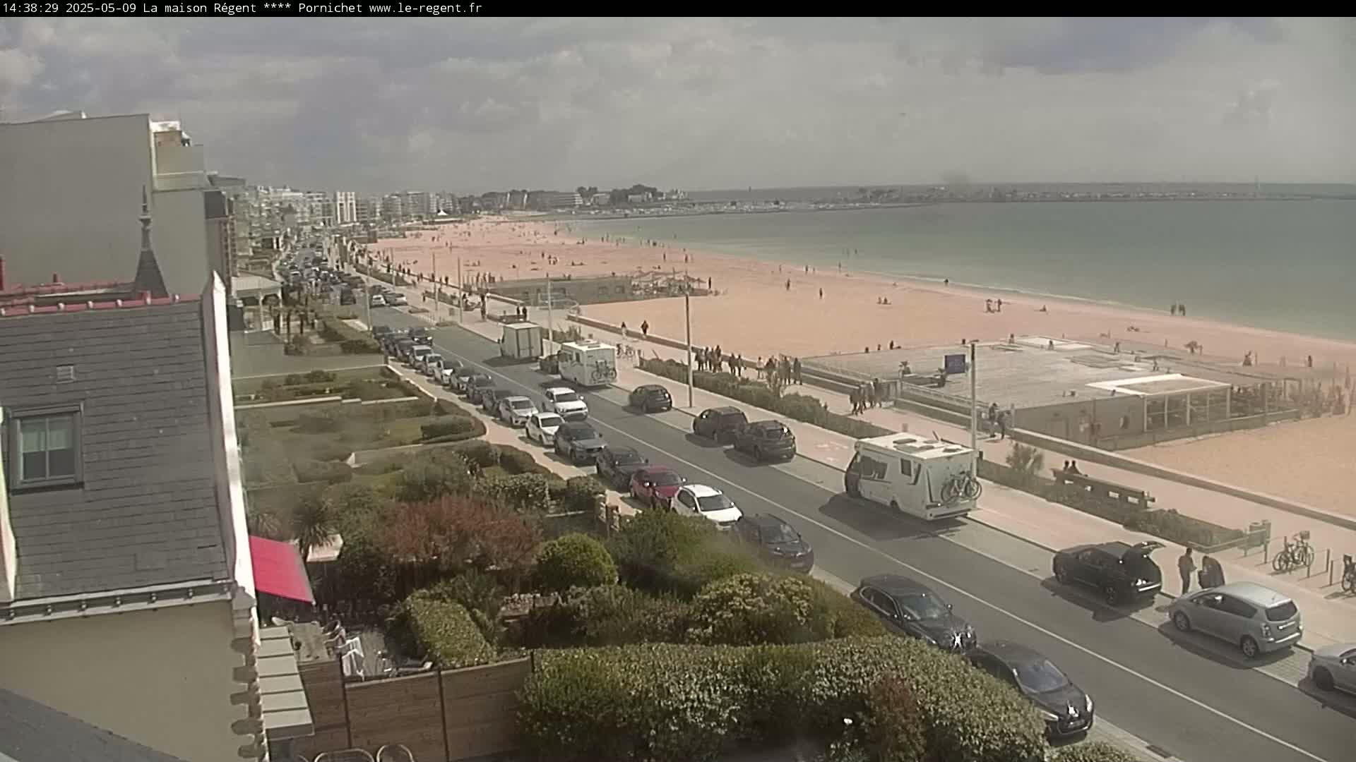 A partly sunny day reveals a beach with many people, a road lined with parked cars, and buildings along a shoreline.