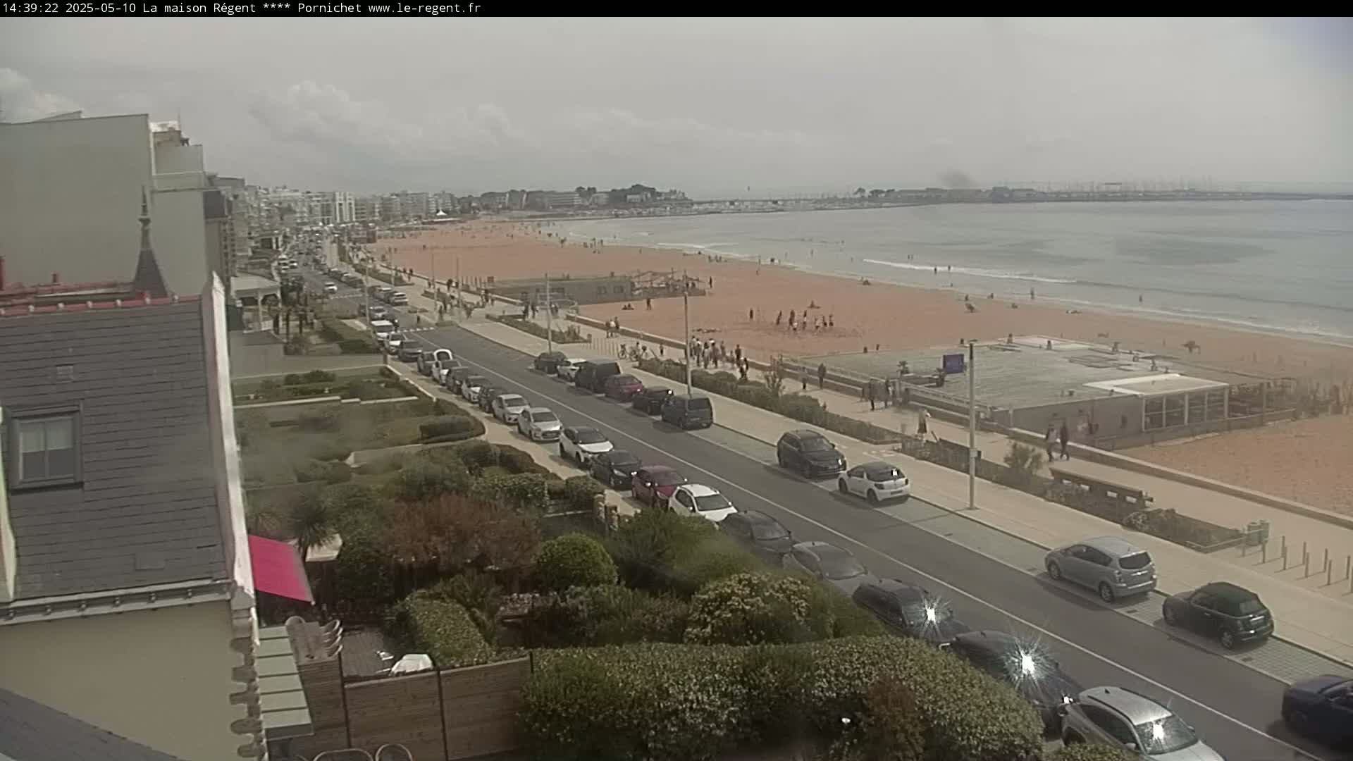 A sandy beach with people sunbathing and swimming is visible from an elevated viewpoint overlooking a seaside road lined with parked cars under an overcast sky.
