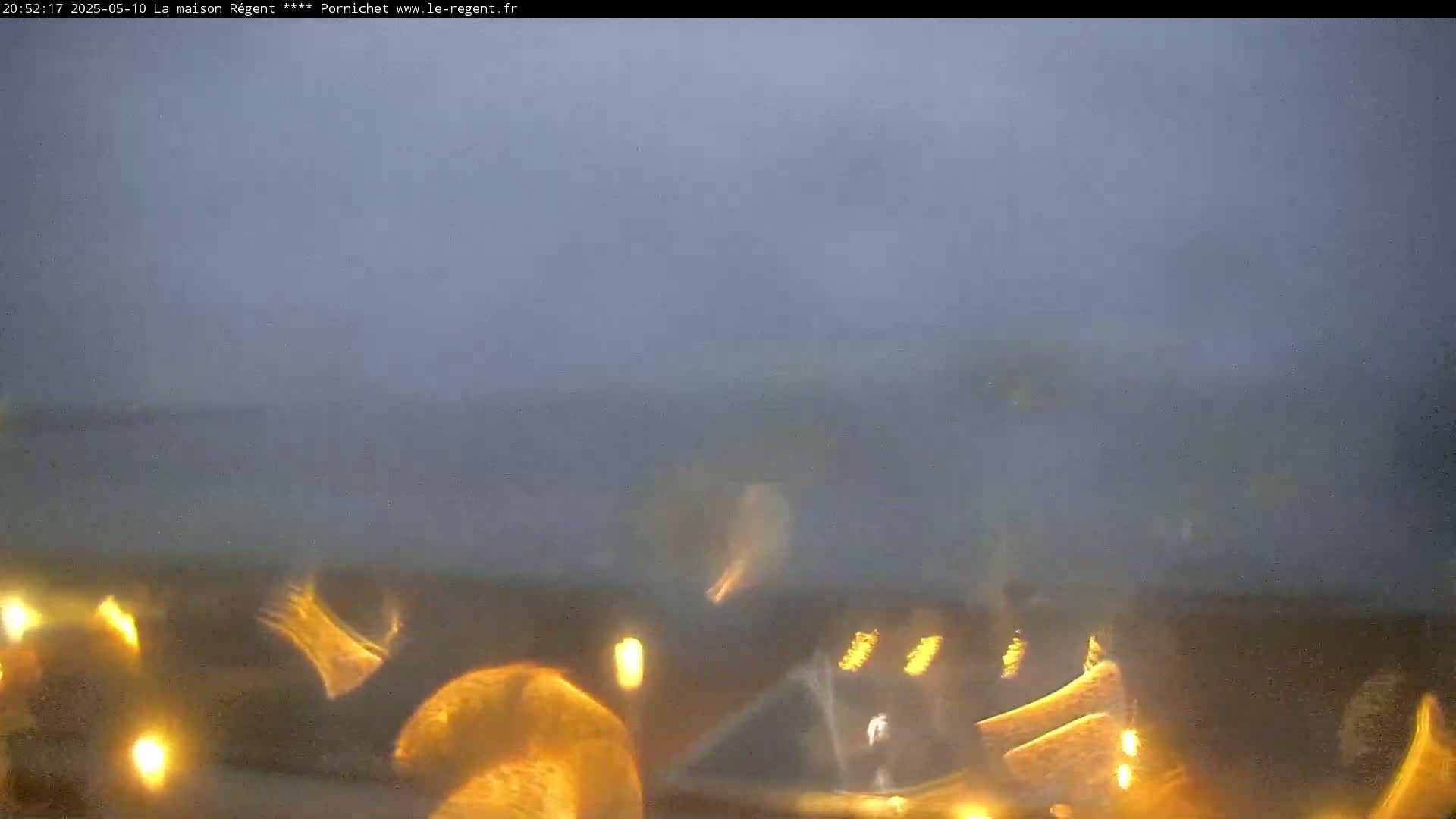 A hazy, dimly lit outdoor scene shows blurry, golden-yellow shapes in the foreground, possibly lights reflecting on a wet surface.