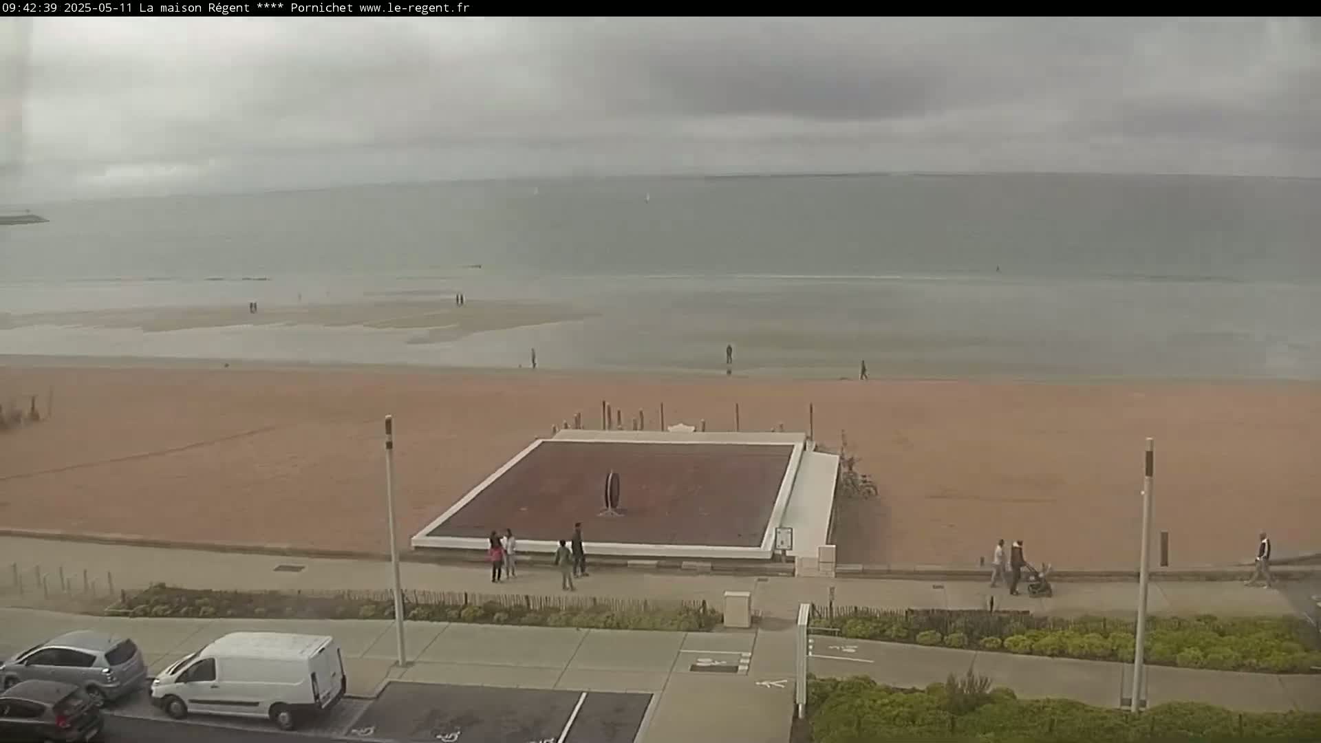 A wide shot shows a mostly empty sandy beach on an overcast day, with a few people walking on the beach and boardwalk near a square structure.