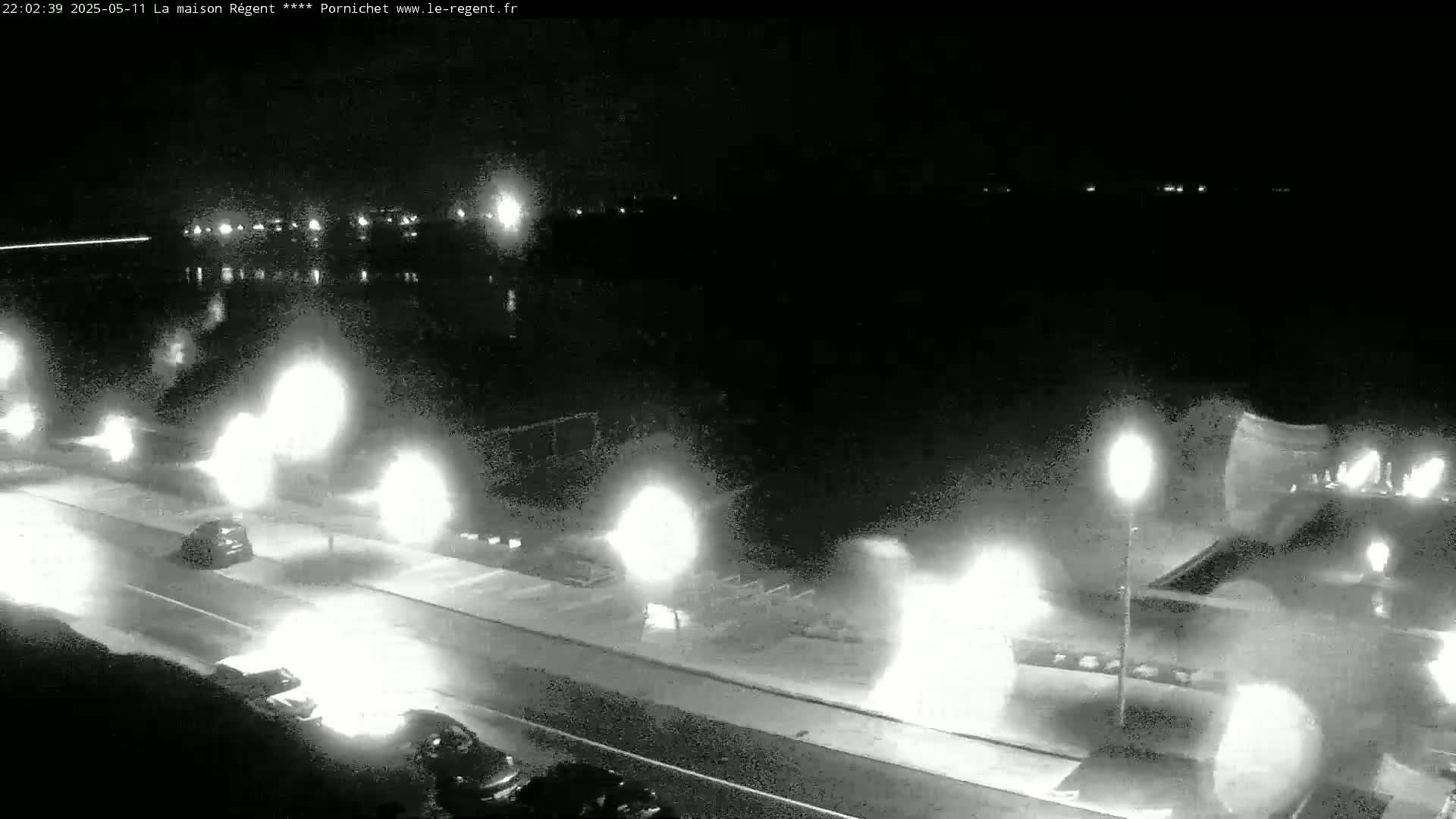 A nighttime, low-light view shows a road beside a body of water with several vehicles driving along the road, their headlights creating bright flares.