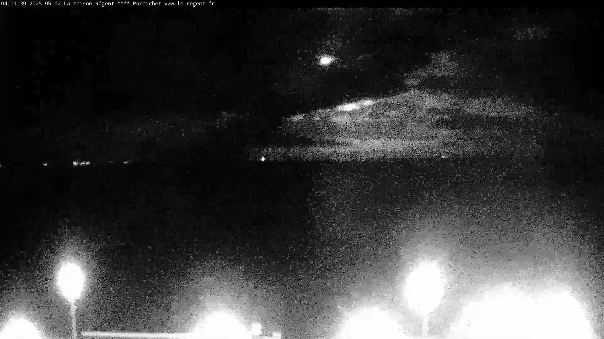 A nighttime, low-light image shows a cloudy sky with distant lights and two bright lights in the bottom corners, likely from ground-level light sources.