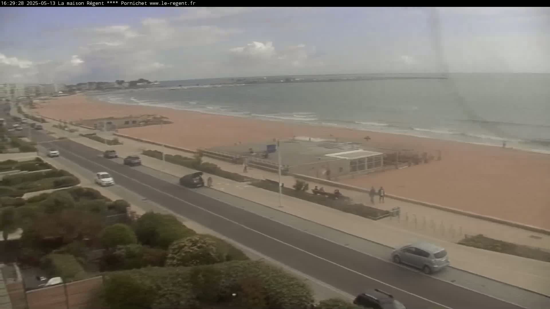 A partly cloudy day shows a sandy beach with calm waves, a road with cars, and people walking along a paved beachfront path.