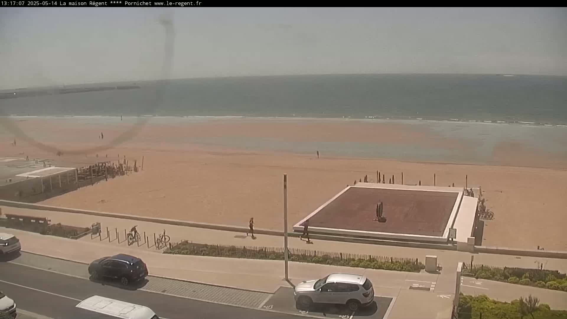 A sandy beach with a calm ocean stretches out under a hazy sky, next to a paved walkway with cars parked nearby, and a few people scattered on the sand and path.