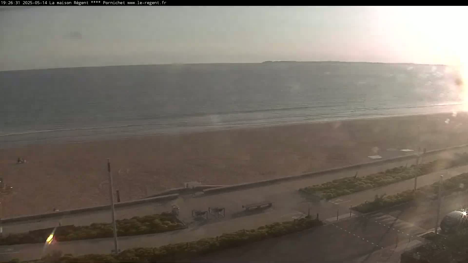 A hazy sunset illuminates a wide sandy beach and calm ocean, overlooking a paved promenade with sparse vegetation.