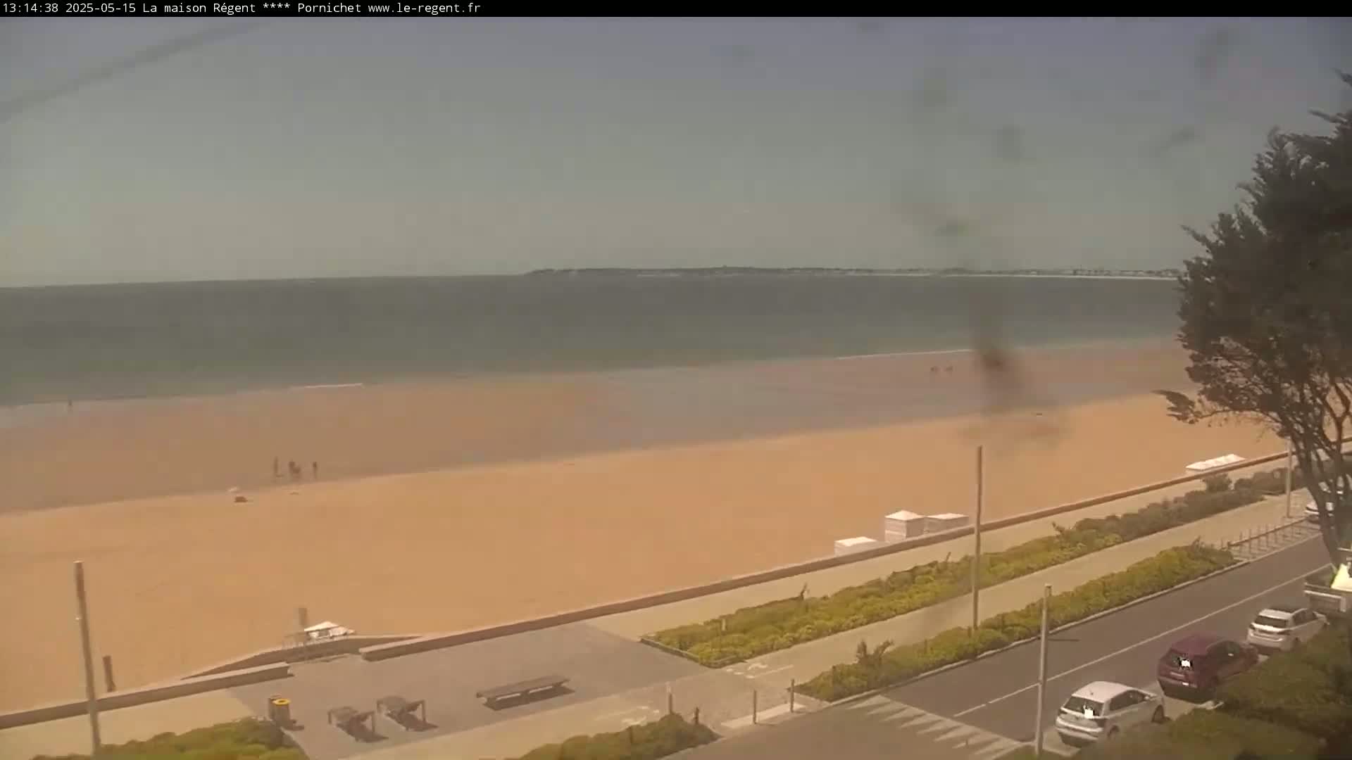 A hazy, sandy beach stretches along a calm ocean under a muted sky, with a few people visible on the sand and cars parked along a nearby road.