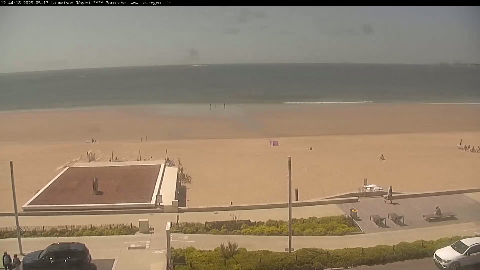 A mostly sunny beach scene shows a large expanse of light-brown sand, a calm ocean, and a few people scattered across the beach and boardwalk.