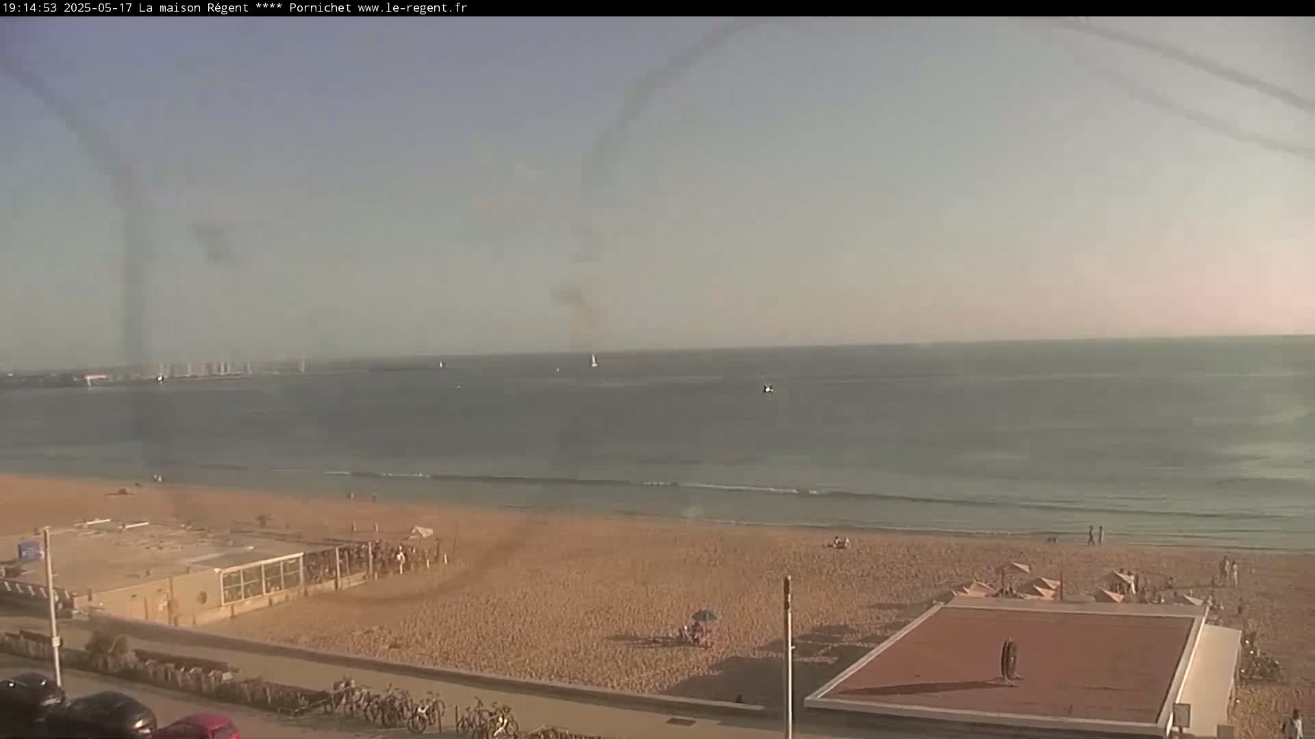 A mostly sunny day at a beach shows calm ocean waves lapping a sandy shore with a few people visible, some buildings and parked bicycles nearby.
