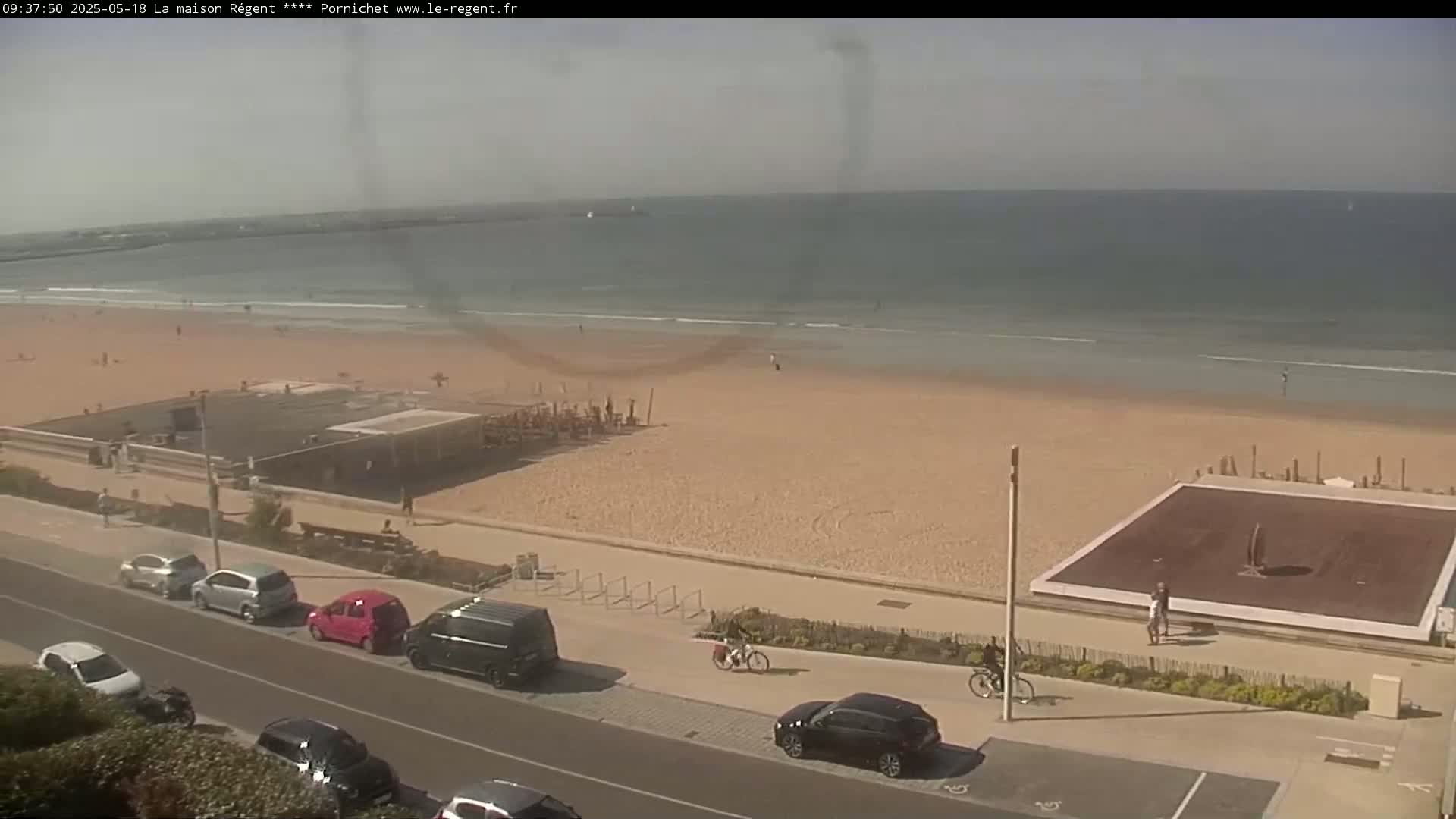 A sandy beach with a calm ocean under a mostly clear sky is viewed from above, showing cars parked along a road next to the beach and several people walking along the shore and boardwalk.