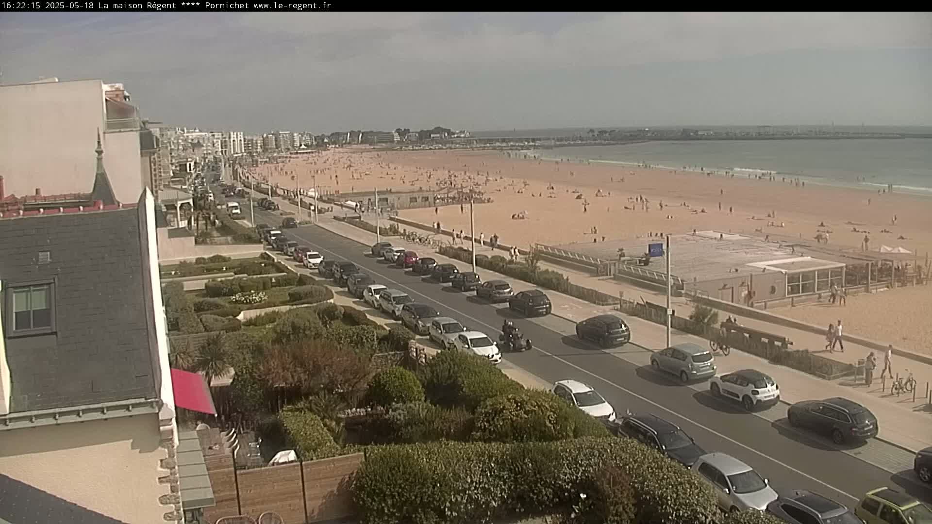 A sunny day at a beach shows many people relaxing on the sand, cars parked along a road next to the beach, and buildings in the background.