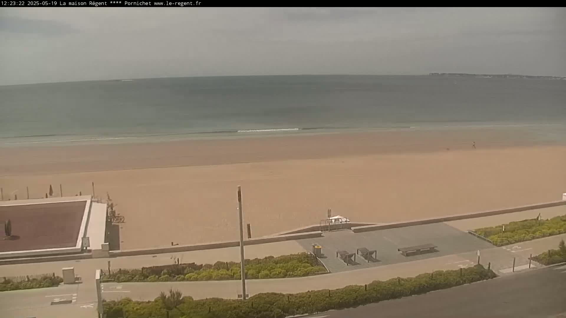 An expansive sandy beach with calm ocean waves stretches out under a mostly cloudy sky, adjacent to a paved walkway lined with greenery and outdoor seating.