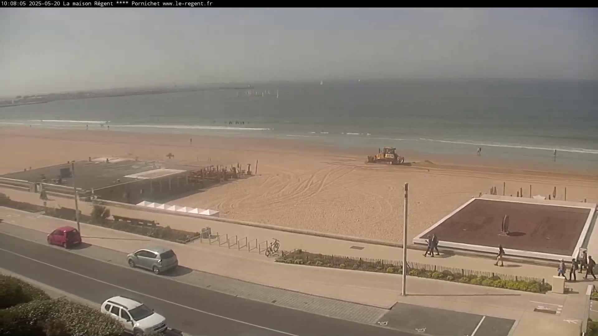 A sandy beach with a bulldozer, a few people, several cars parked along a road, and a calm ocean under a clear sky.