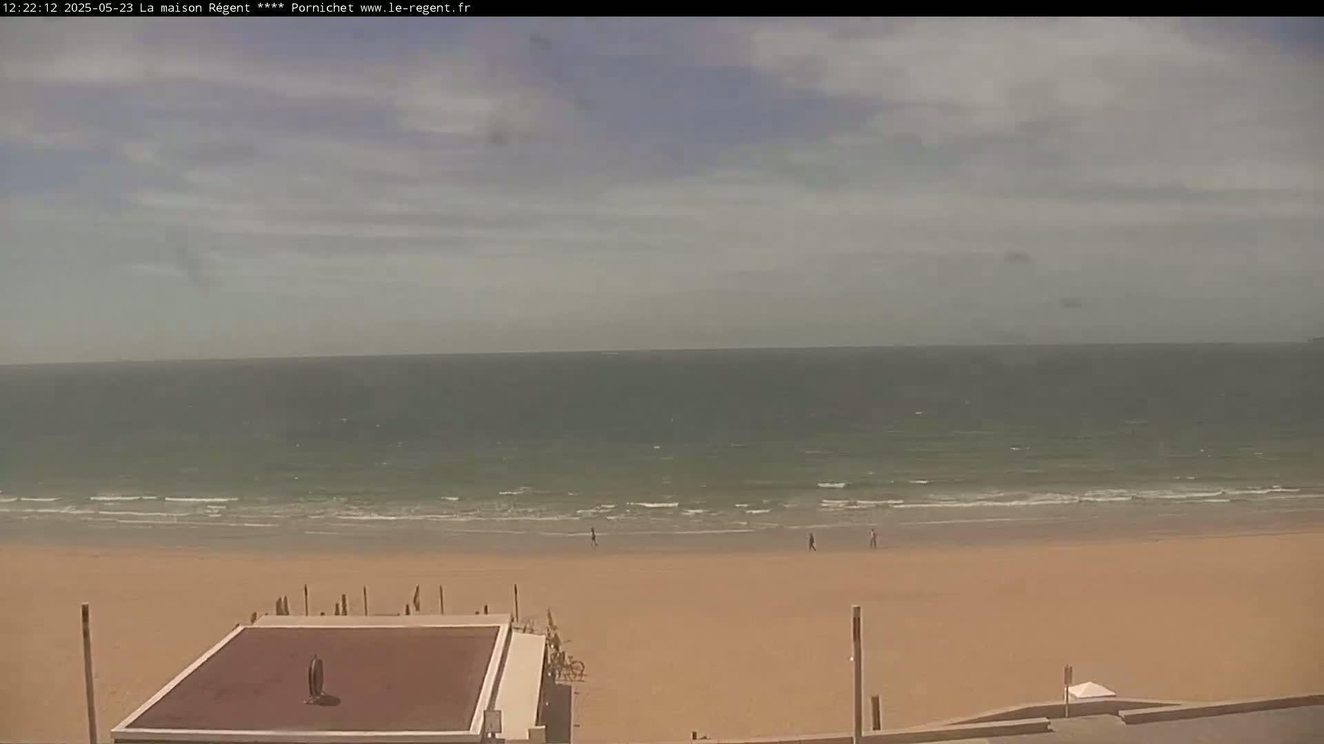 A sandy beach with a few people walking on it, gently lapping waves, and a mostly cloudy sky is visible from an elevated vantage point.