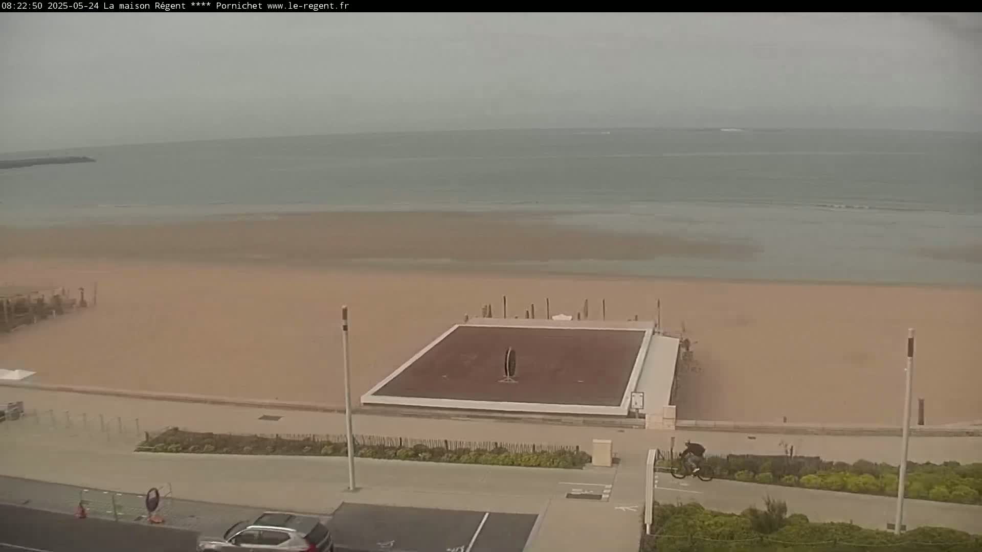An overcast day at a beach shows a calm ocean, a wide sandy beach, a paved boardwalk, a raised flat square area, and a person riding a bicycle.
