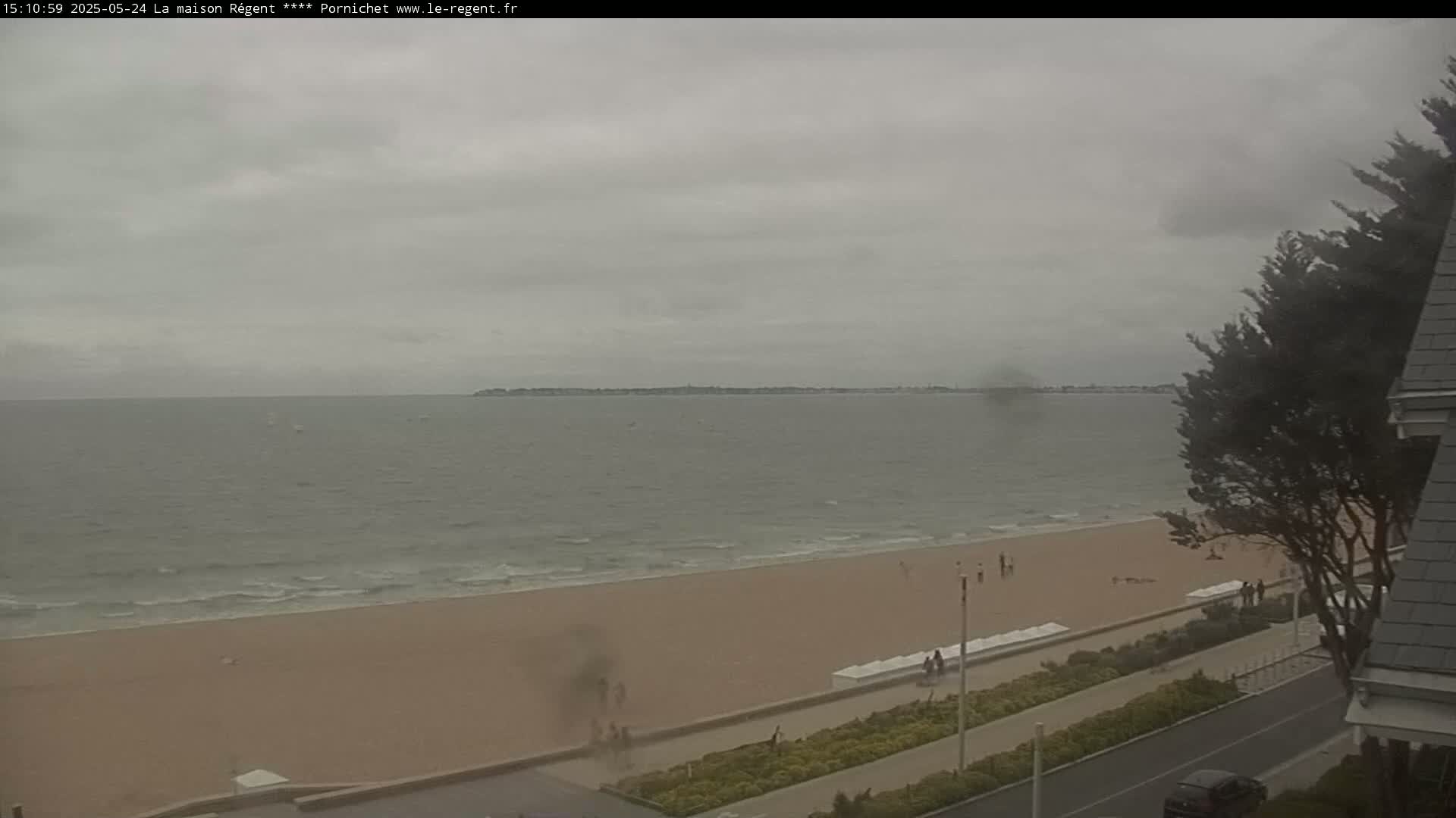 A sandy beach stretches along a calm, gray sea under an overcast sky, with a few people visible on the shore and a tree partially obscuring the view from a high vantage point.