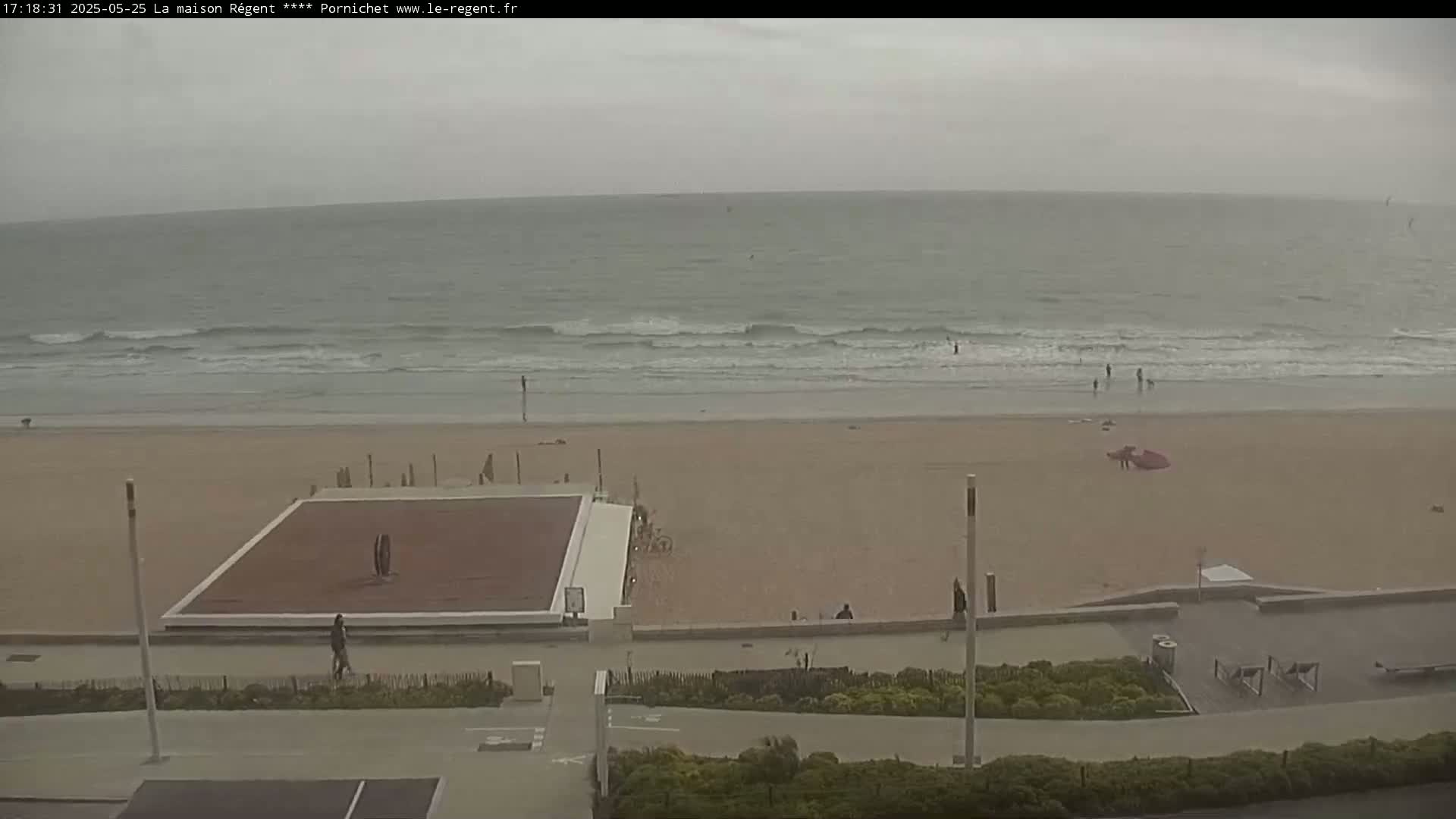 An overcast day shows a beach with small waves, a few people on the sand and in the water, and an empty paved plaza area nearby.