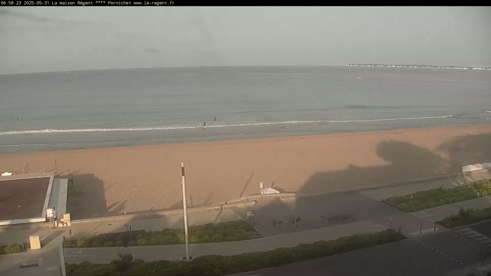 An overcast day at a beach shows calm waves lapping a wide sandy shore, with a paved walkway and sparse vegetation visible beyond the sand.