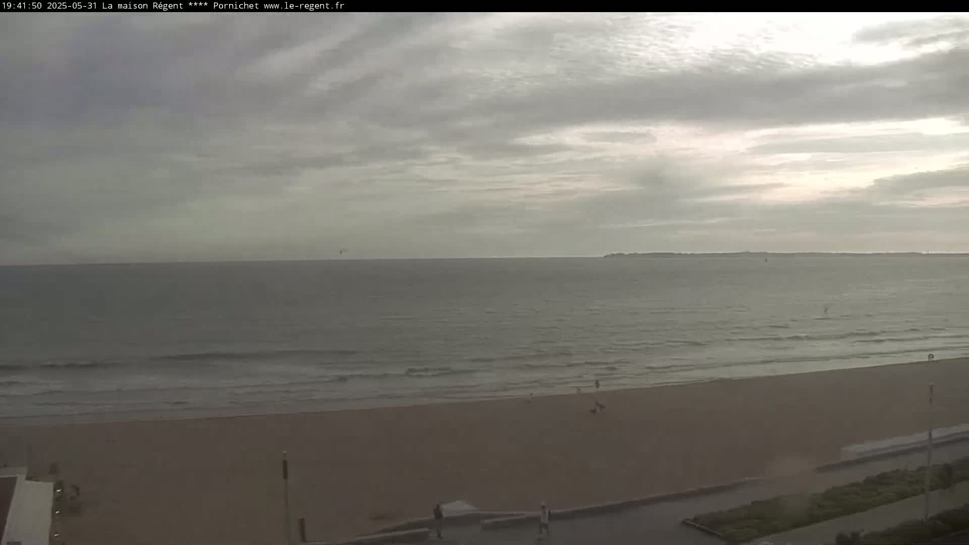 An overcast sky hangs over a wide sandy beach and calm ocean with a few people visible on the shore.