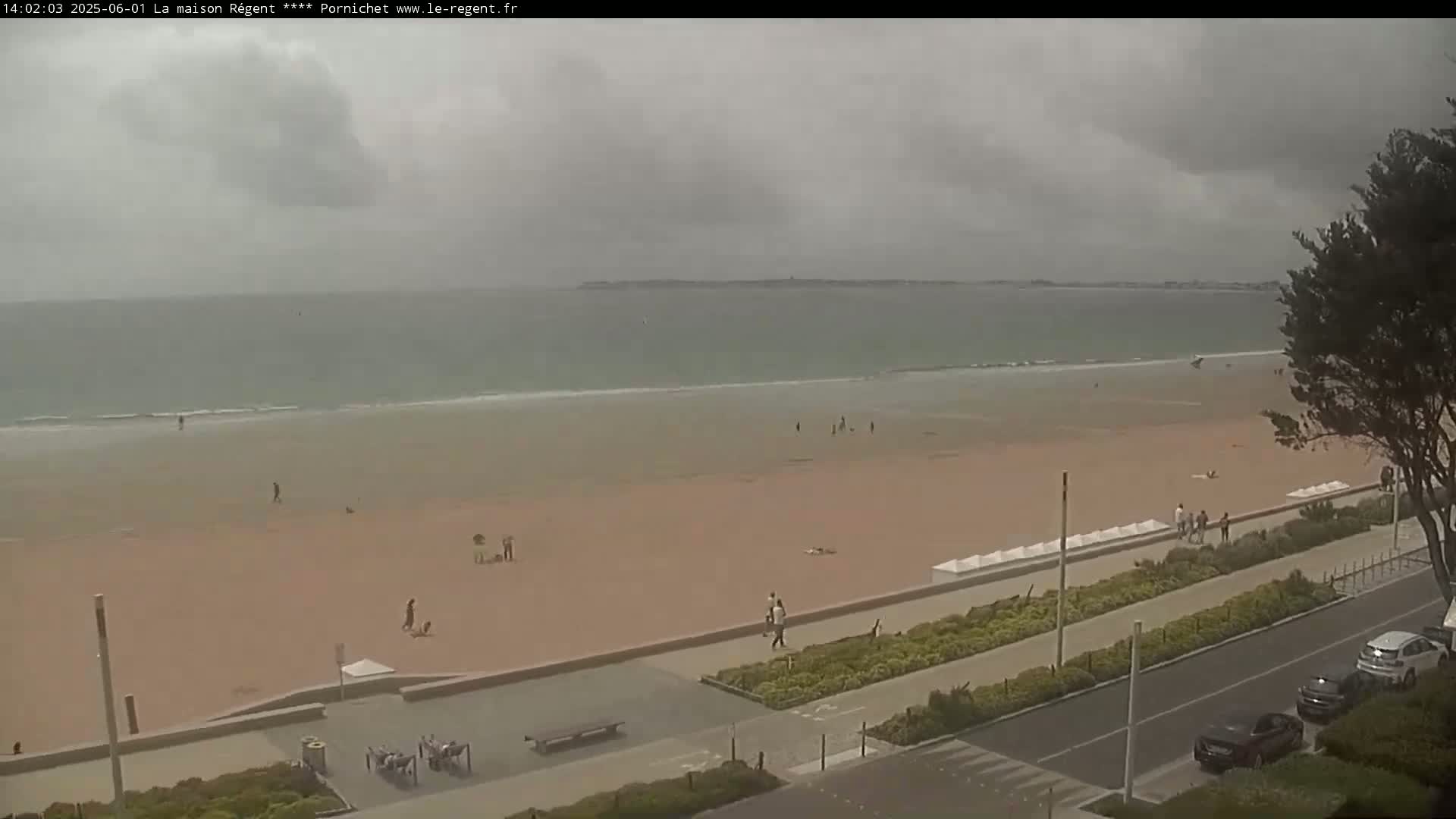 A mostly overcast day shows a sandy beach with a few people scattered across it, next to a paved walkway and a street with parked cars.