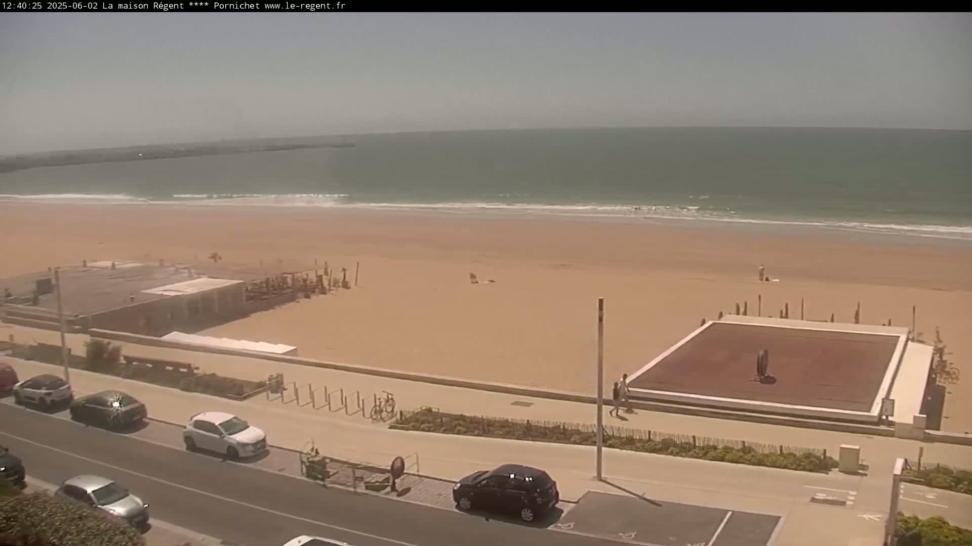 An overhead view shows a sandy beach with calm ocean waves under a bright, sunny sky, a boardwalk with parked cars and a building near the beach.