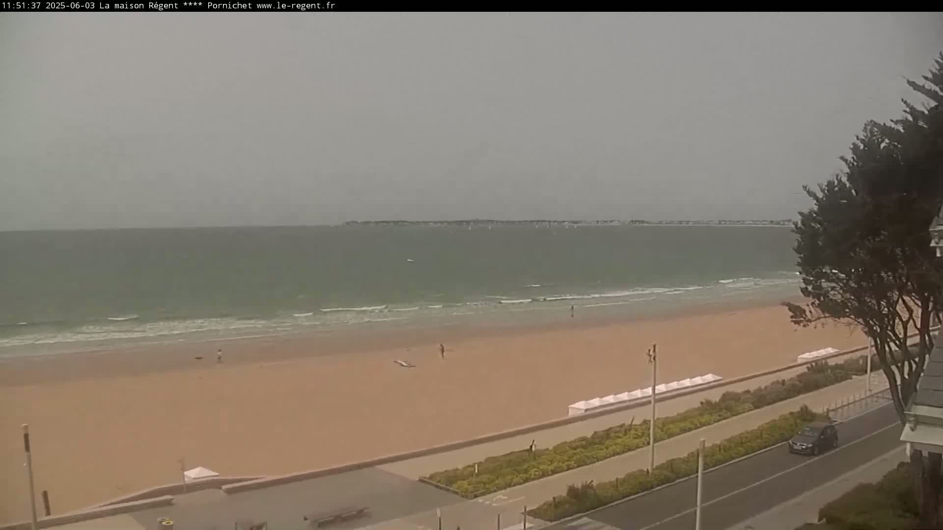 A mostly cloudy day shows a wide sandy beach with a few people and a calm ocean with small waves, viewed from an elevated position overlooking a roadway and some vegetation.