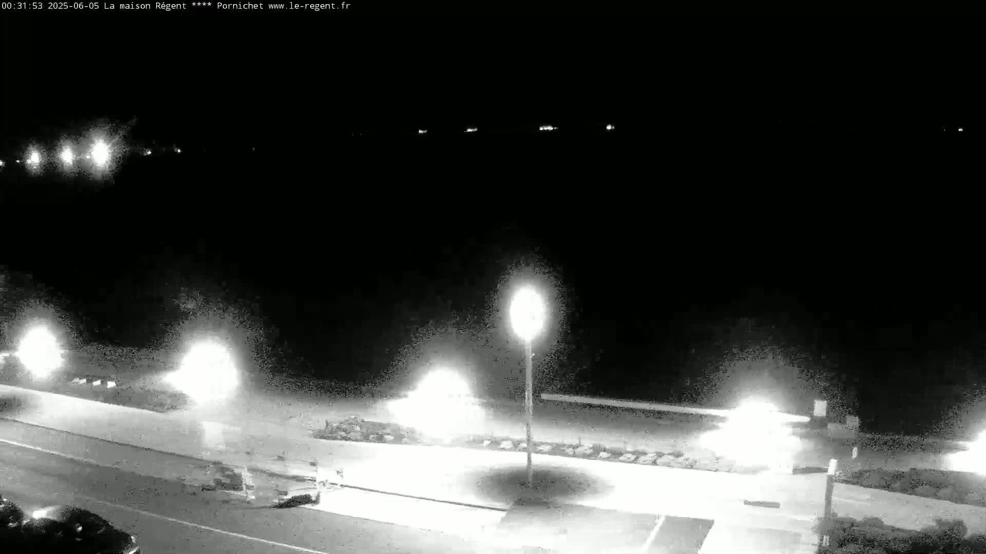 A nighttime, black and white view of a dark, paved road next to a beach, illuminated by bright streetlights.