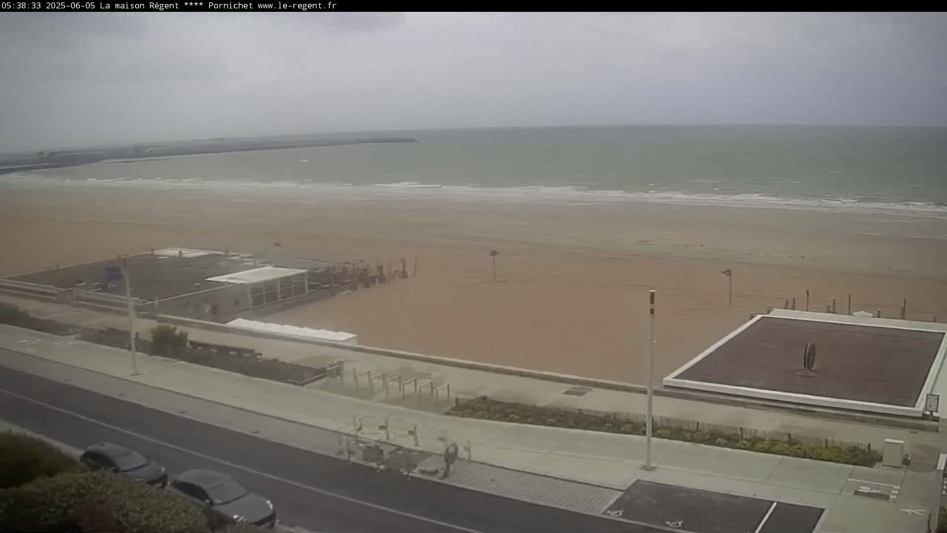 An overcast day at a beach shows a wide sandy shore, calm ocean waves, beachfront buildings, and a paved walkway with parked cars.