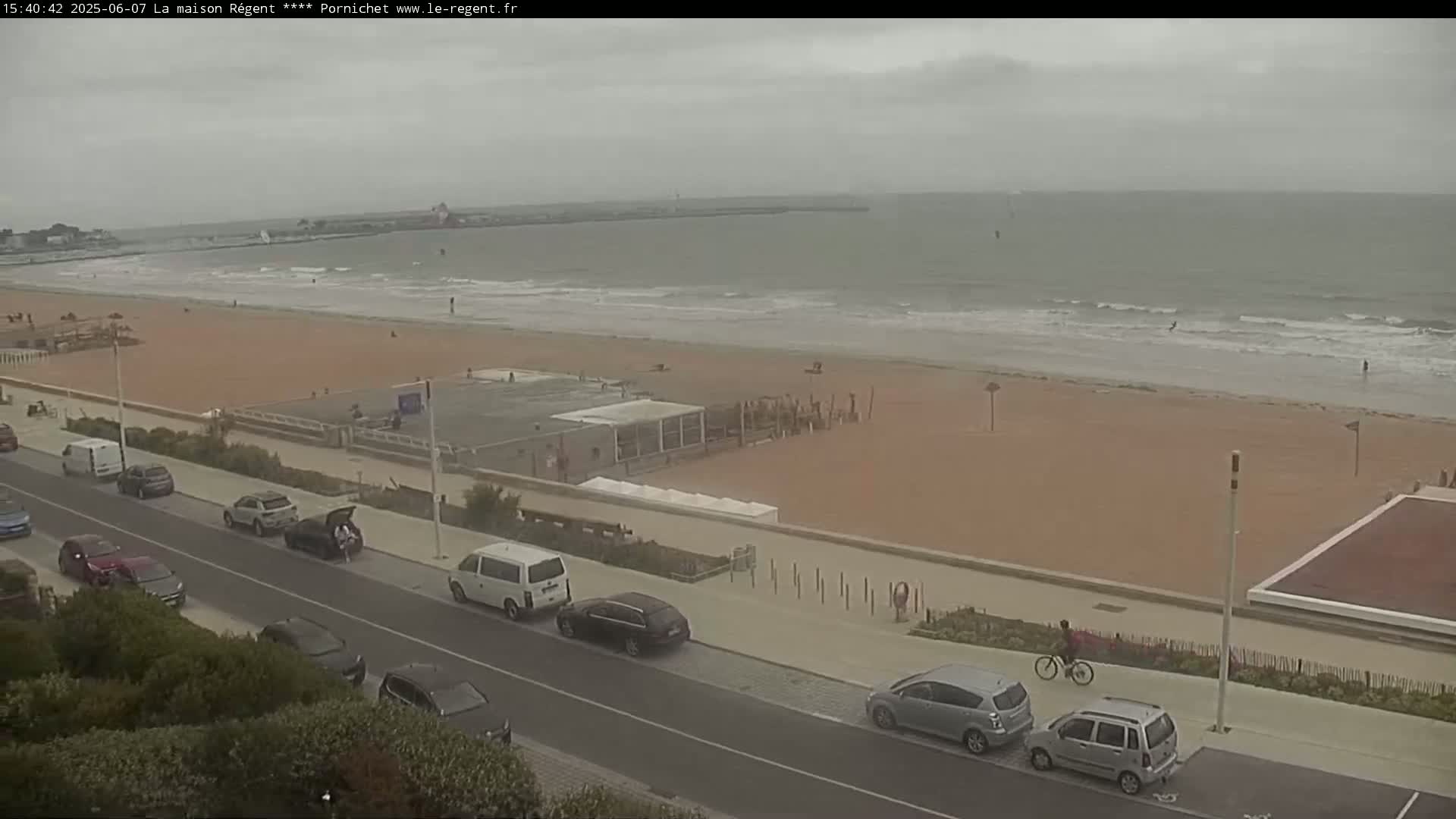 A beach with overcast skies and small waves is seen from an elevated vantage point, showing a road with parked cars beside it, and a few people are visible on the beach and the road.