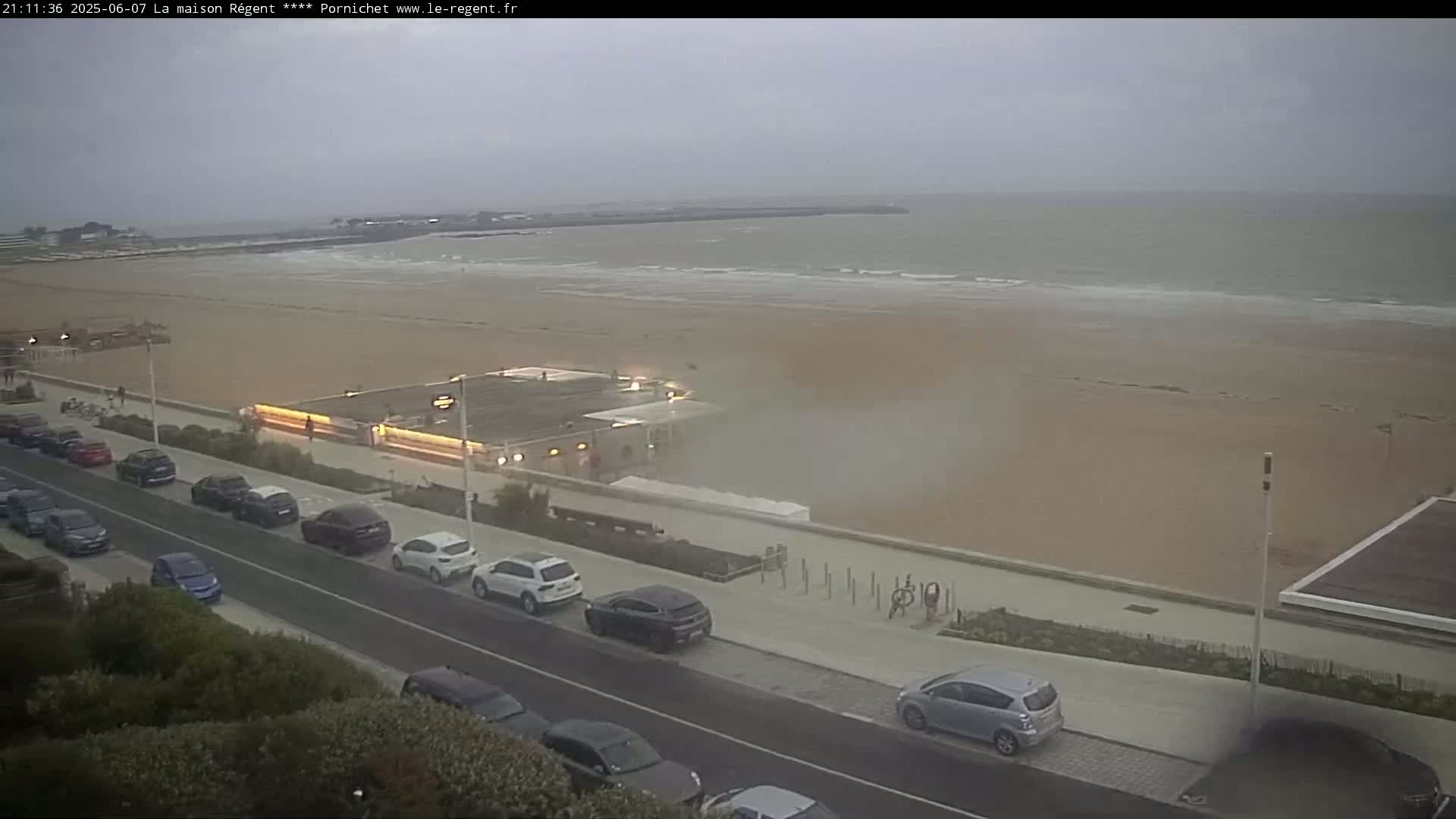 A beach with a row of parked cars along a paved walkway next to it, under an overcast sky.