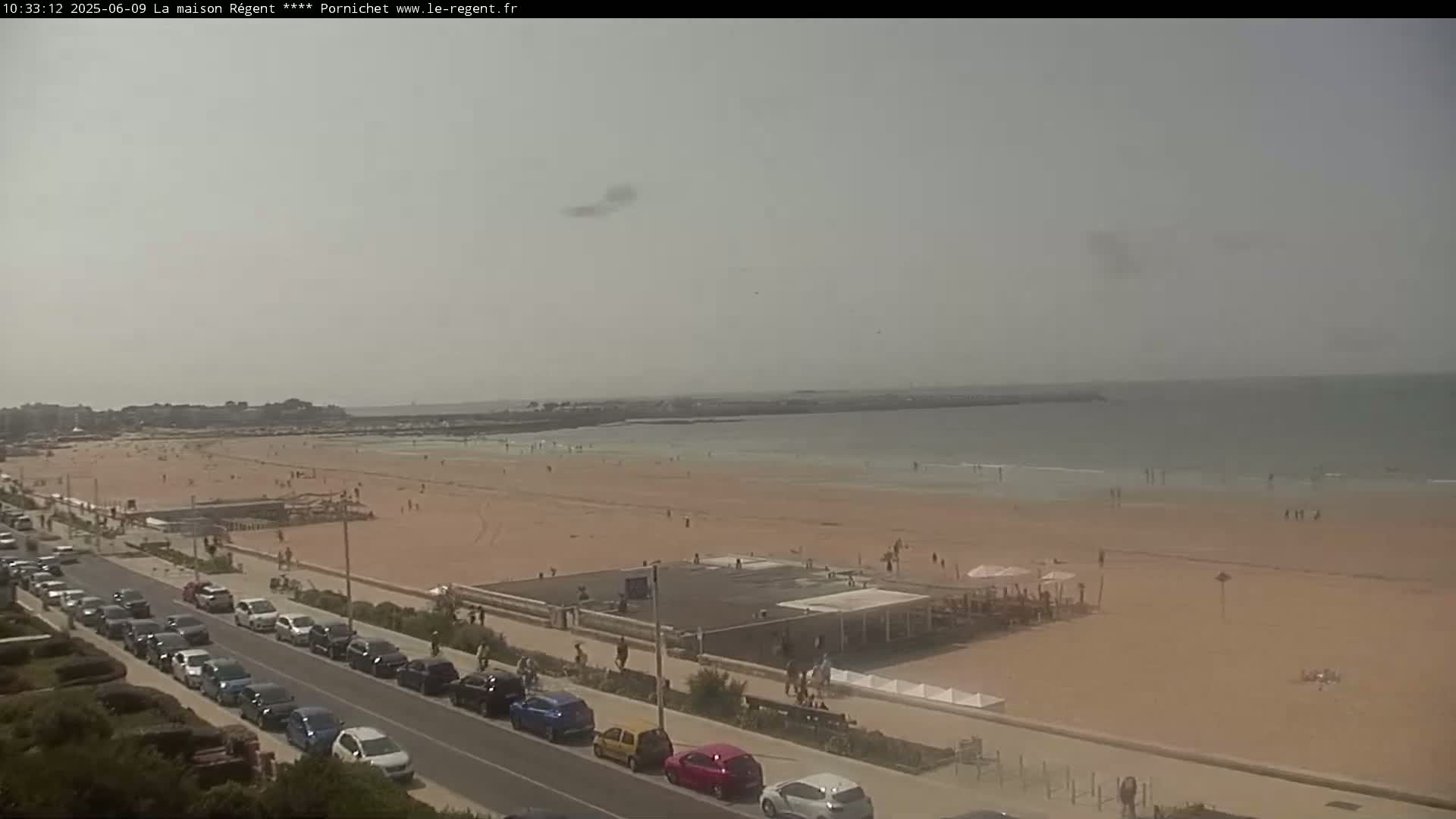 A long sandy beach with scattered people stretches to a calm sea under a hazy sky, a road lined with parked cars runs parallel to the shore.