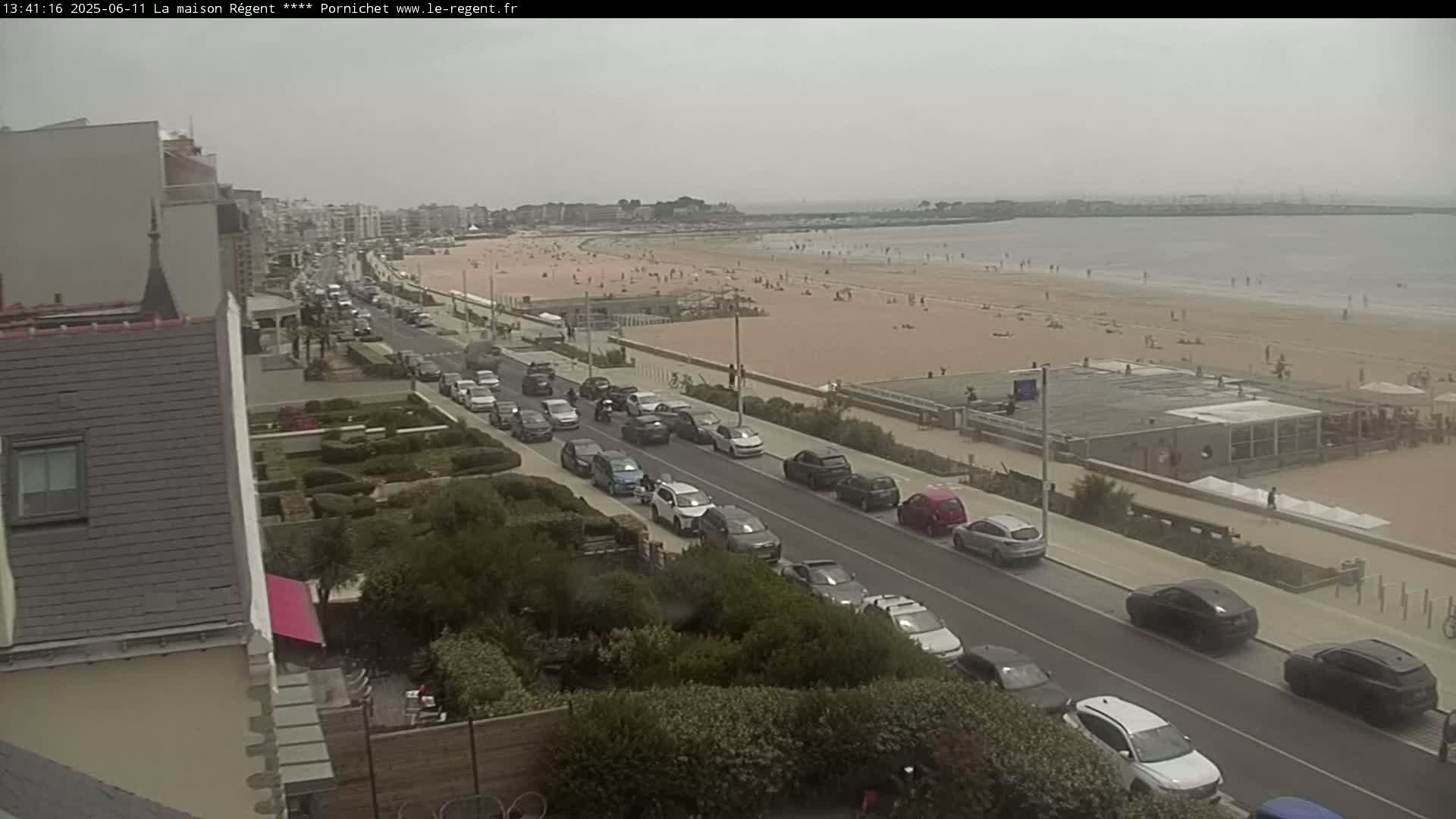 A beach with many people on it is visible from a street with parked cars, under an overcast sky.