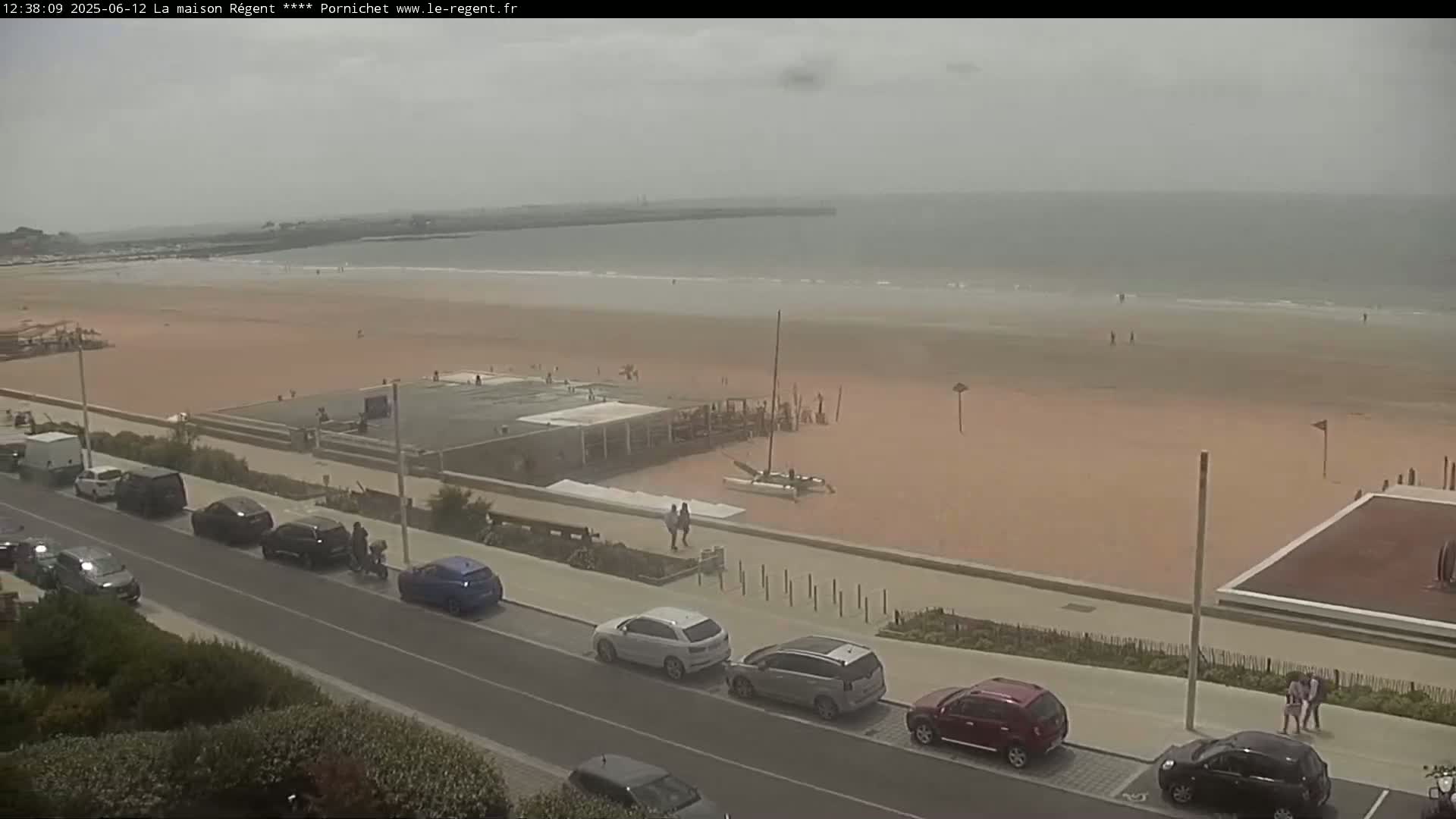 A sandy beach with a few people and cars parked along a road next to it, under an overcast sky.