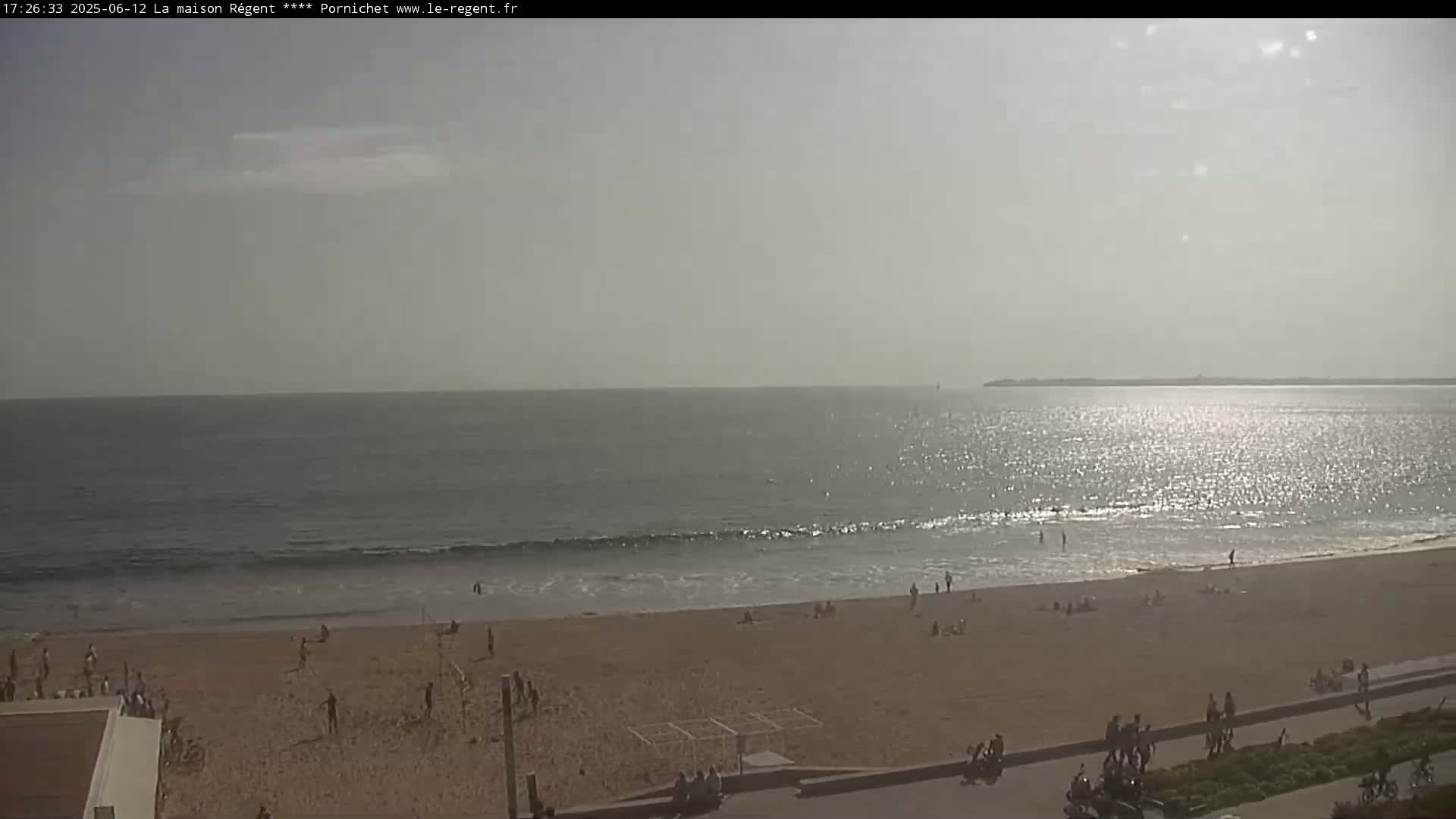 A mostly sunny day at a beach shows a calm ocean with small waves, and many people are relaxing or playing on the sandy shore.
