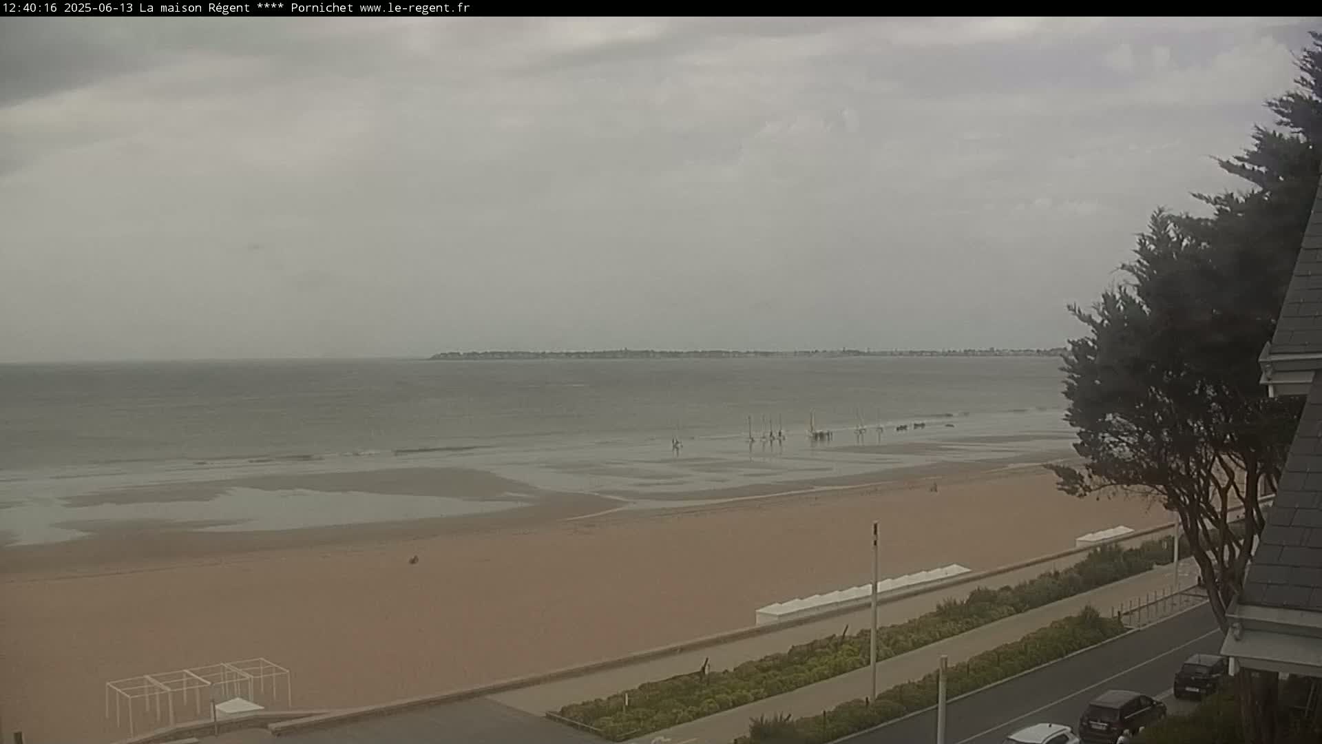 A wide sandy beach stretches to a calm, gray sea under an overcast sky, with a few sailboats visible in the distance and cars parked along a road bordering the beach.