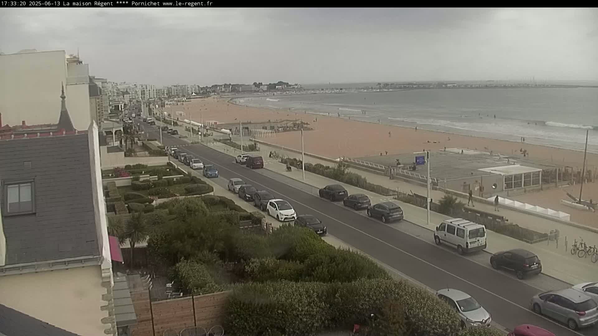 A seaside street with parked cars overlooks a sandy beach with a few people on it under an overcast sky.