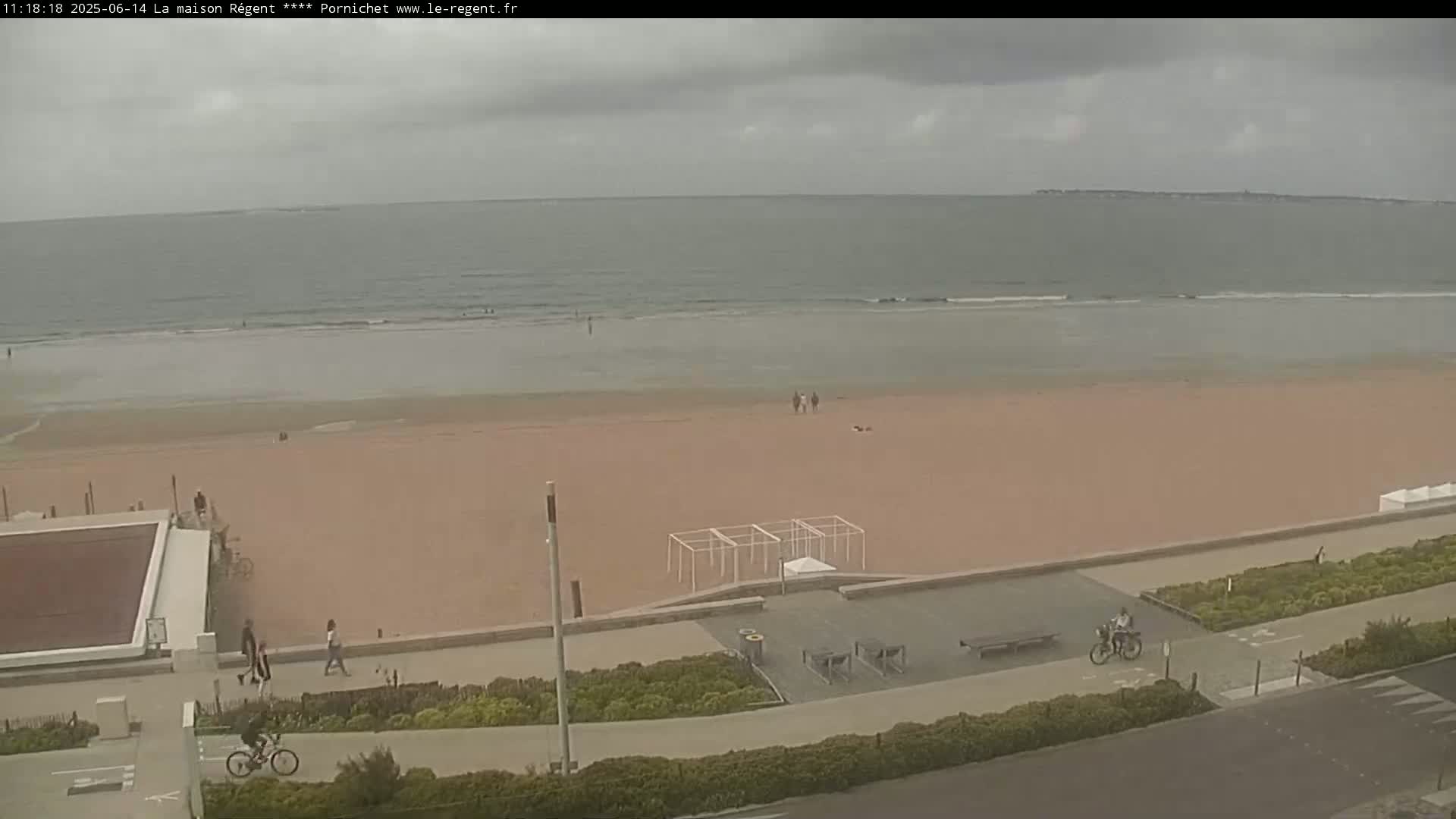 A mostly cloudy day shows a wide sandy beach with a calm ocean, a few people walking and biking, and some beachside structures.